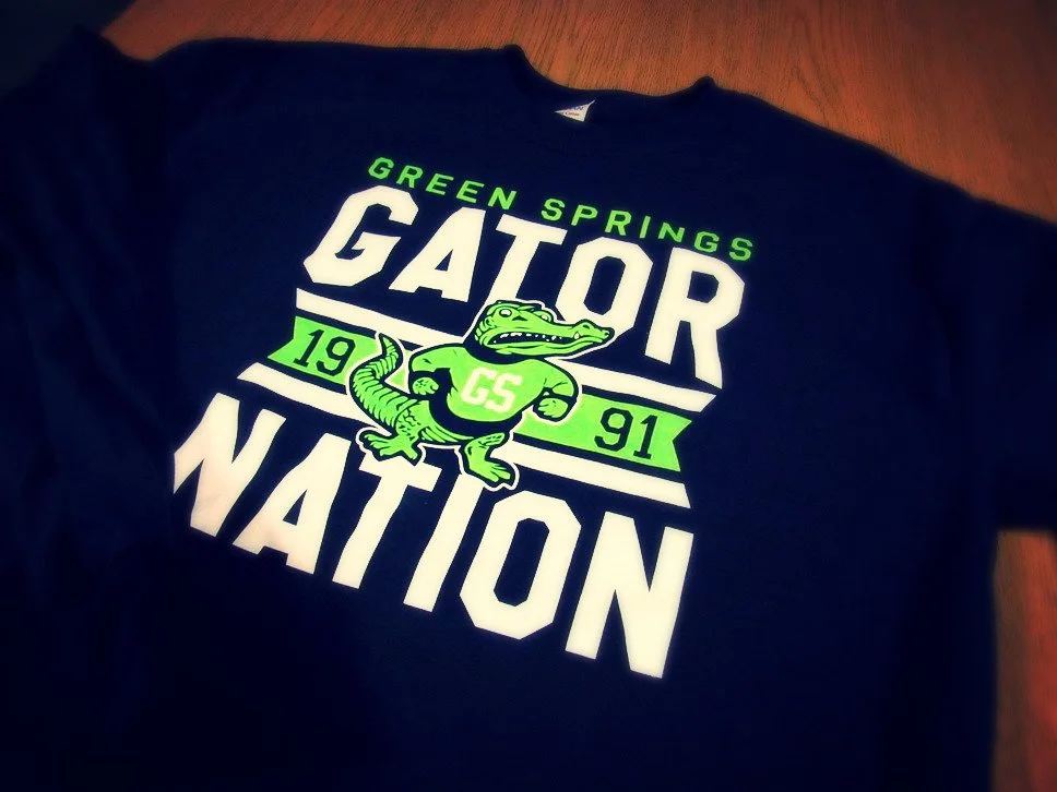 Black T-shirt with green and white text that reads "Green Springs Gator Nation" and features a cartoon alligator wearing a jersey with the initials "GS" and the year "1991" on it.