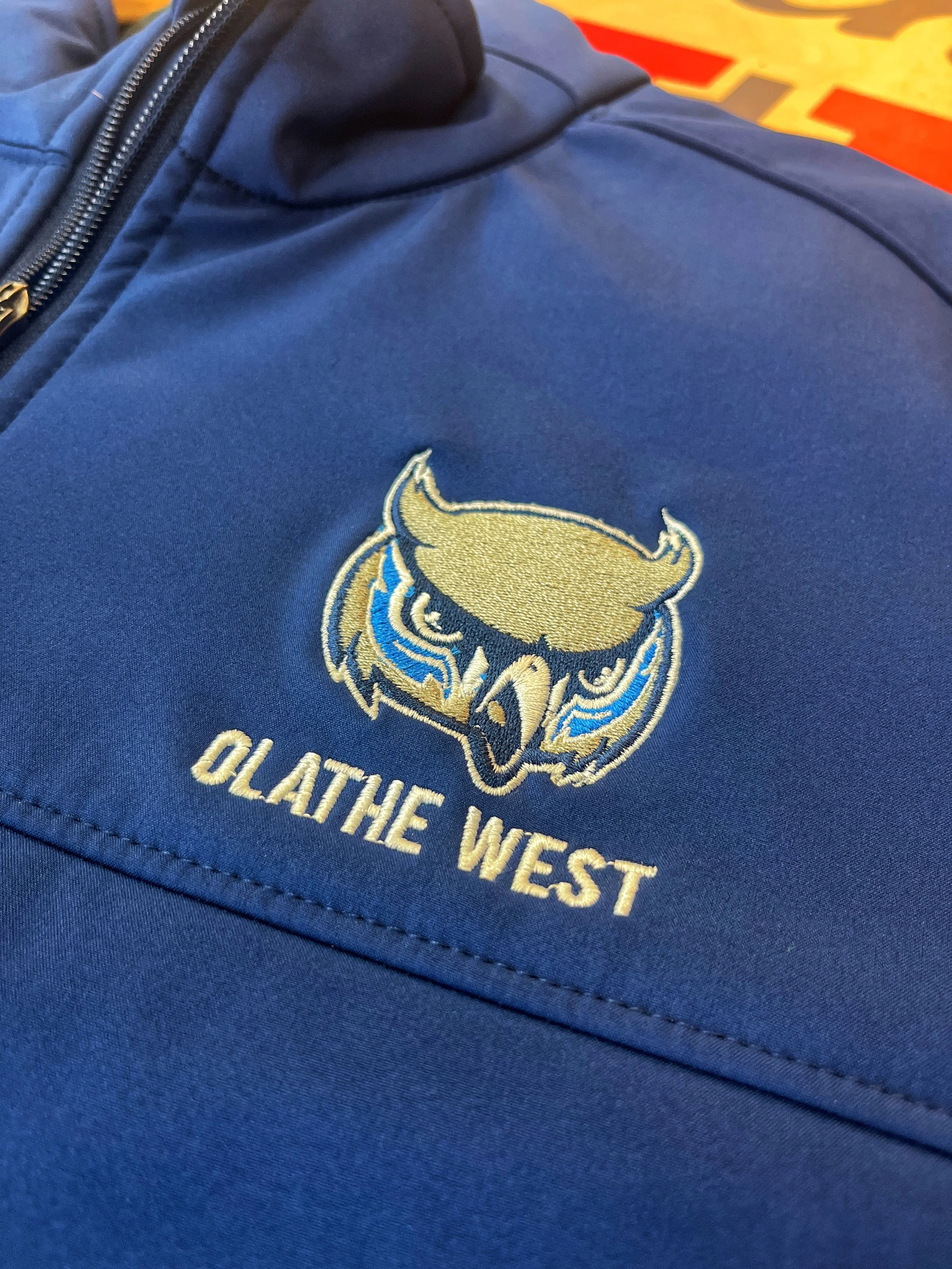 Close-up of a gold Olathe West owl logo embroidered on navy apparel.