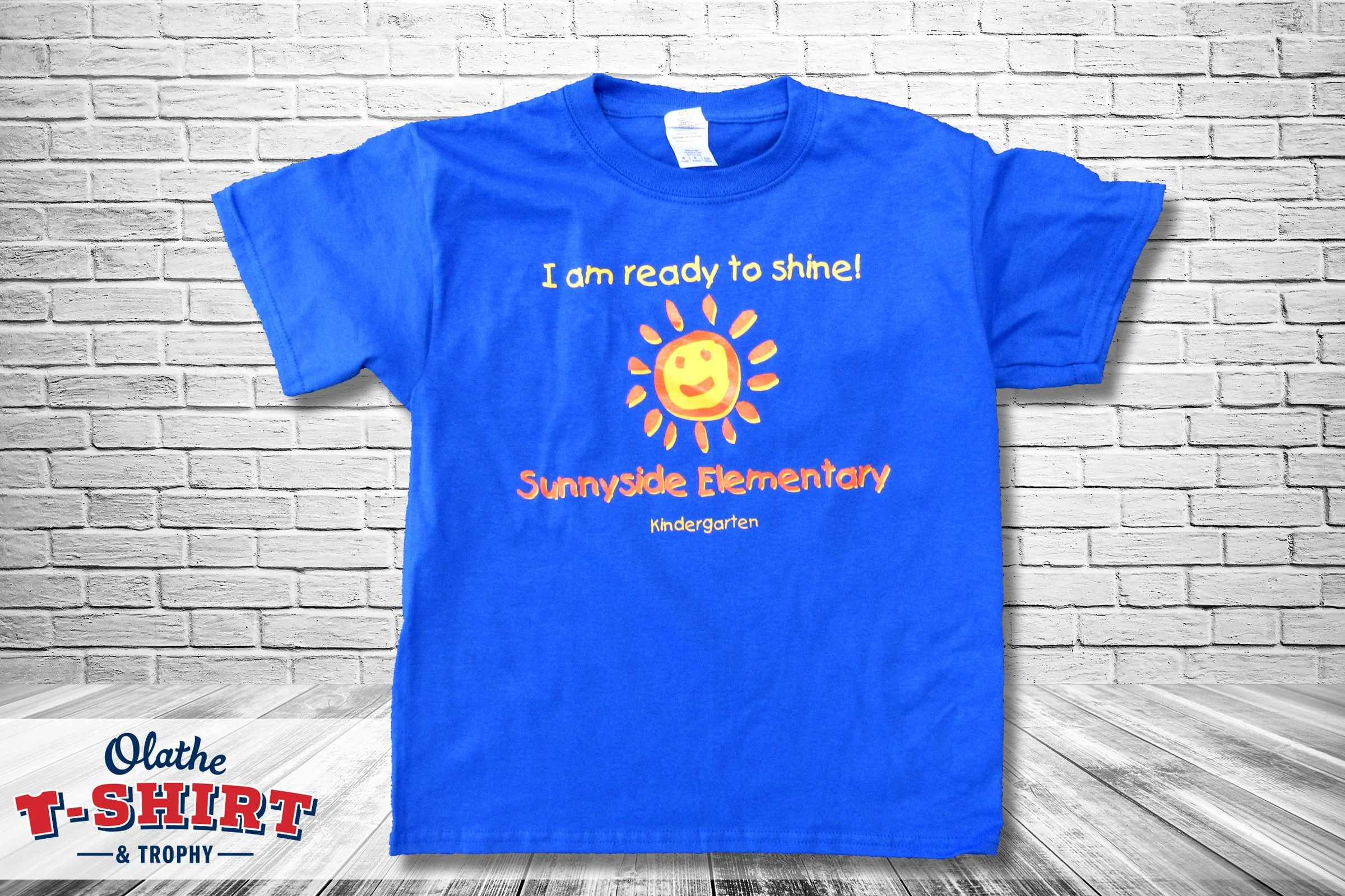 Blue T-shirt hanging against a white brick wall. The T-shirt has a cheerful sun with a smiling face and the text 'I am ready to shine!' above it. The bottom of the shirt features the words 'Sunnyside Elementary' and 'Kindergarten'.