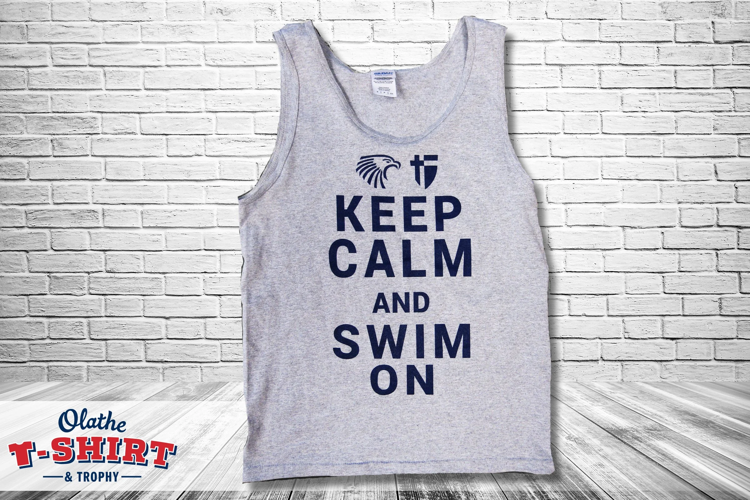 Gray sleeveless T-shirt hanging against a white brick wall with the text 'KEEP CALM AND SWIM ON' printed on it in dark blue. Part of a logo or text in red and blue is visible at the bottom left corner.