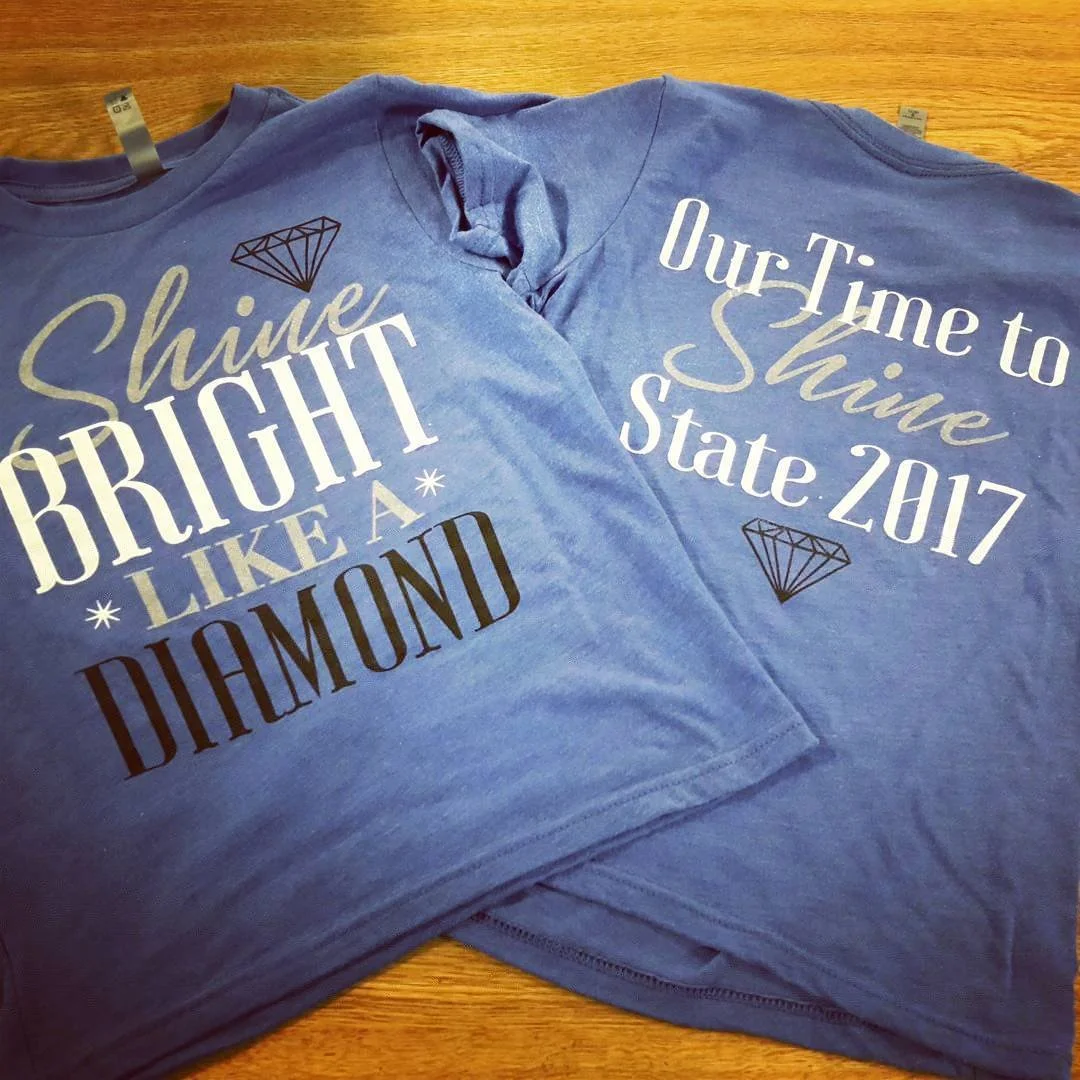 Two blue T-shirts with white and black text and diamond graphics, laid flat on a wooden surface. One shirt says, 'Shine Bright like a Diamond' and the other says, 'Our Time to Shine State 2017.'