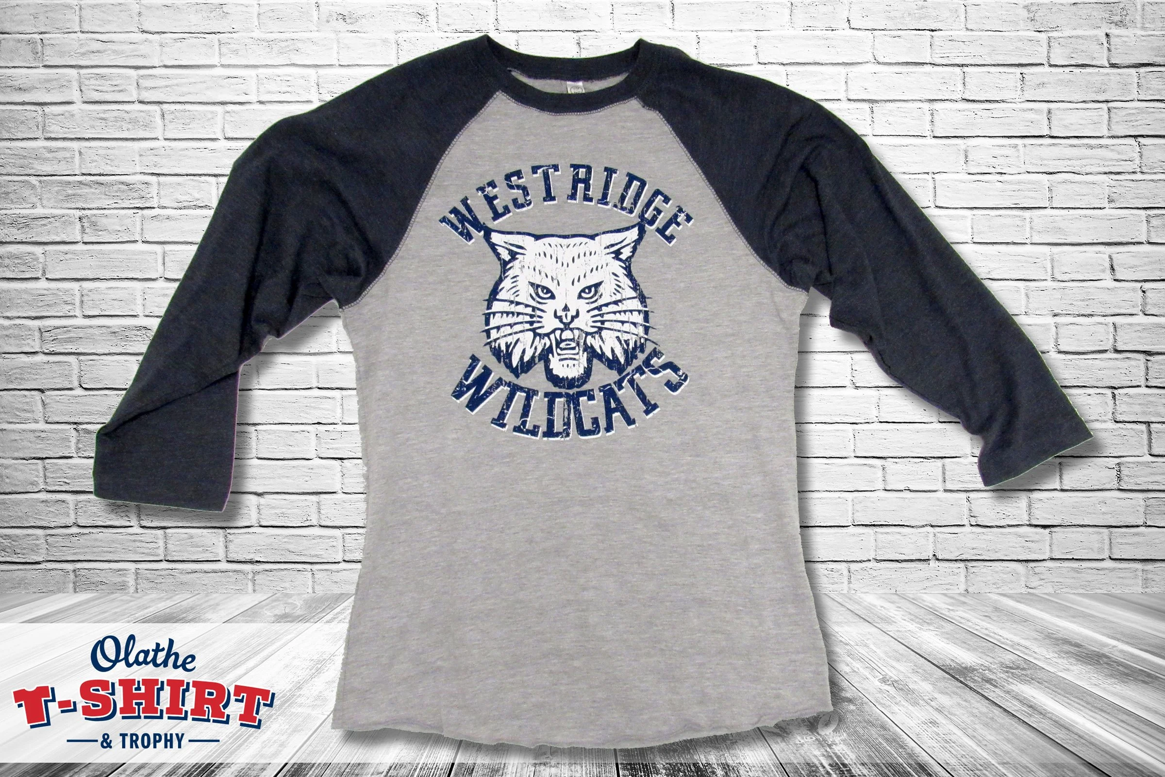 Gray and black baseball-style shirt with a wildcat mascot, displayed against a white brick wall.
