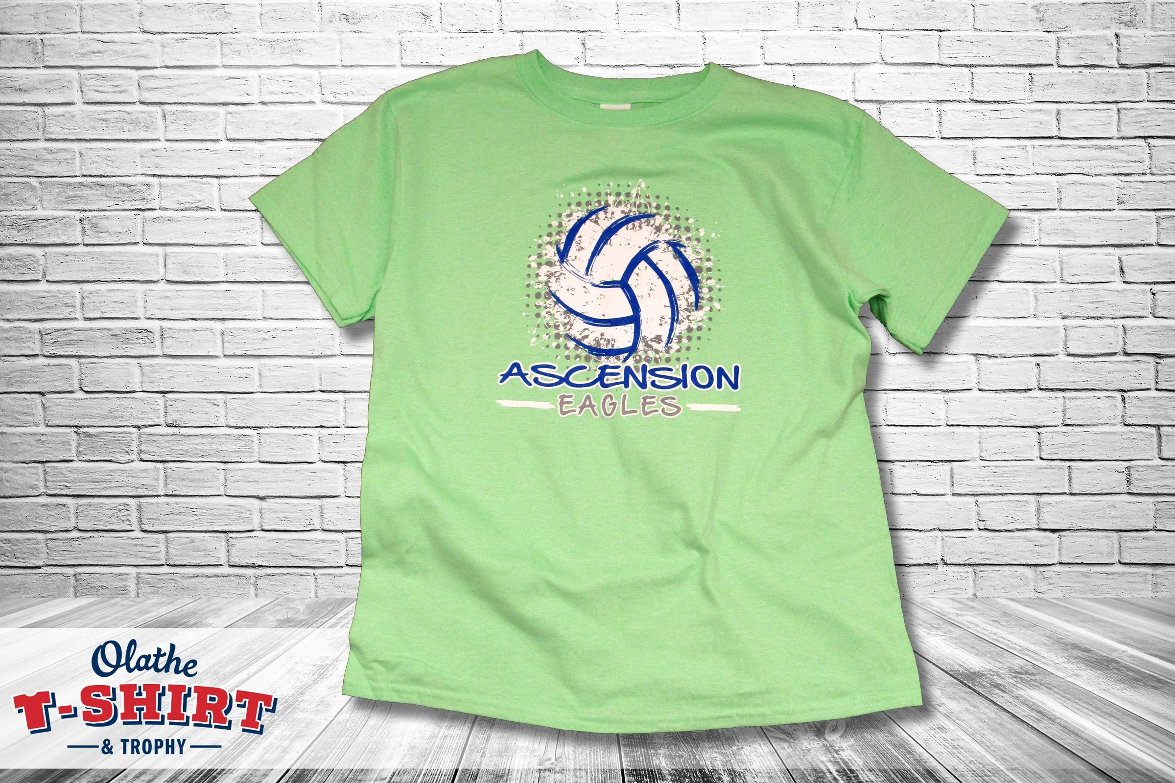 A green T-shirt with a graphic of a volleyball and the words "Ascension Eagles" on the front, hanging against a white brick wall.