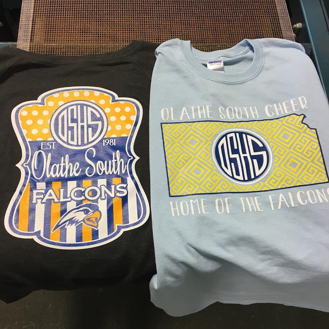 Two T-shirts, one black and one light blue, with Olathe South Falcon logos and slogans. The black shirt has a colorful crest with the Falcon logo, and the blue shirt has a yellow geometric design and the same Falcon logo.
