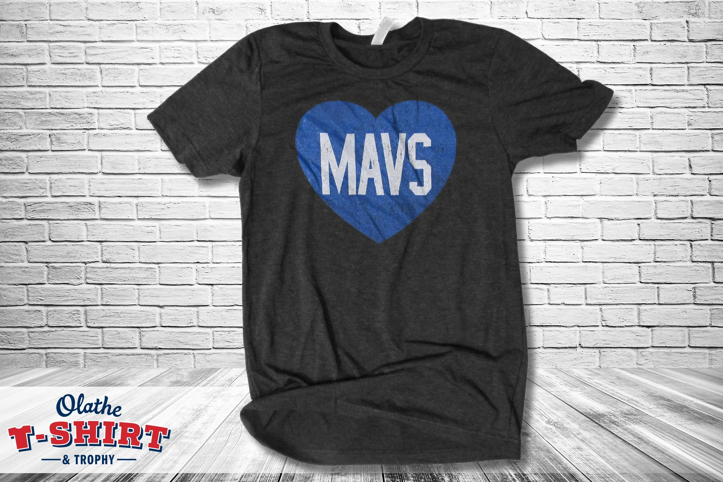 A black T-shirt hanging on a white brick wall with a blue heart and white text that reads 'MAVS' in the center.
