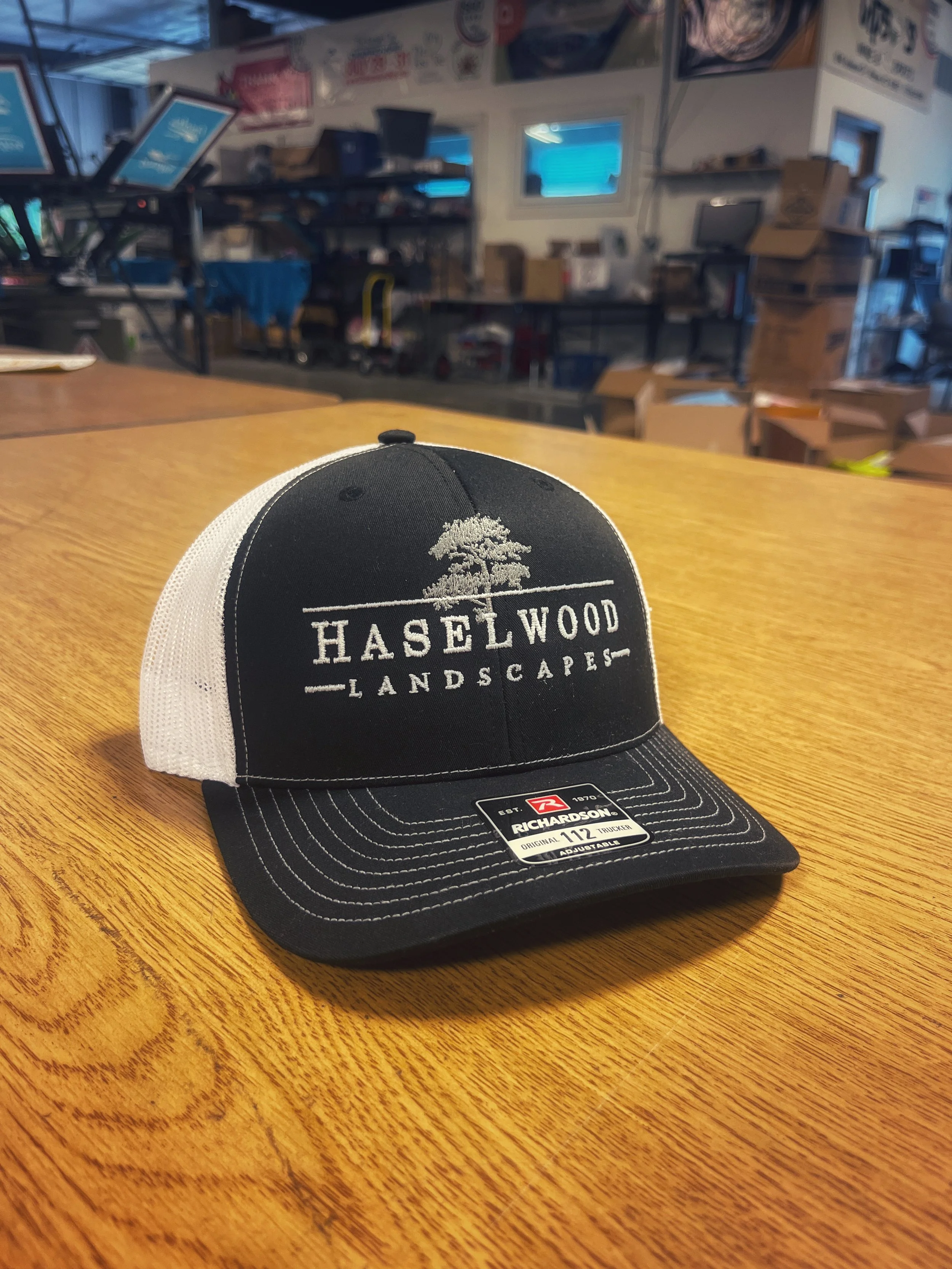 Black and white trucker cap with an embroidered landscaping company logo and tree graphic.