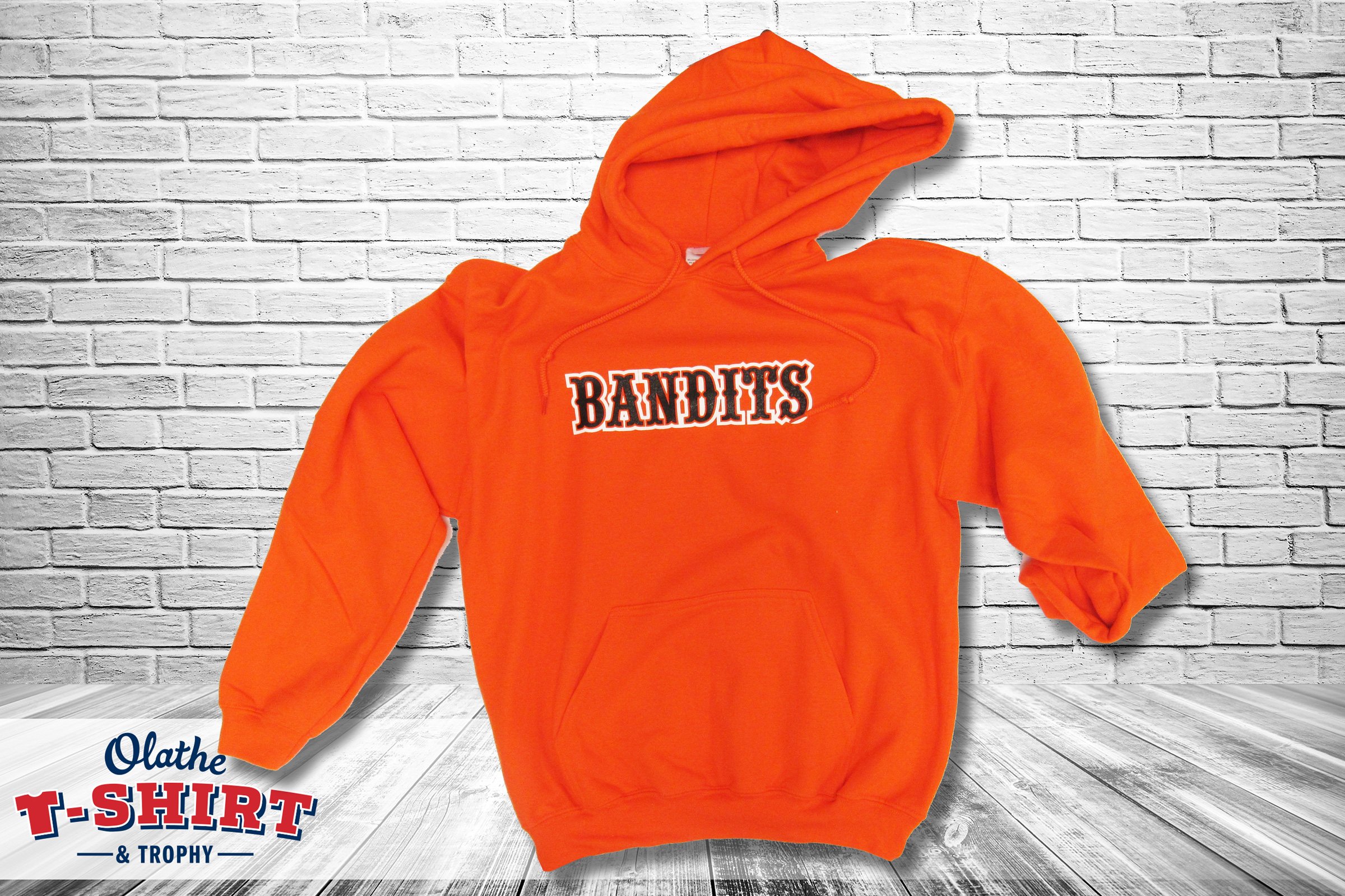 An orange hoodie with the words "Bandits" printed on the chest in bold black and white lettering, hanging against a white brick wall.
