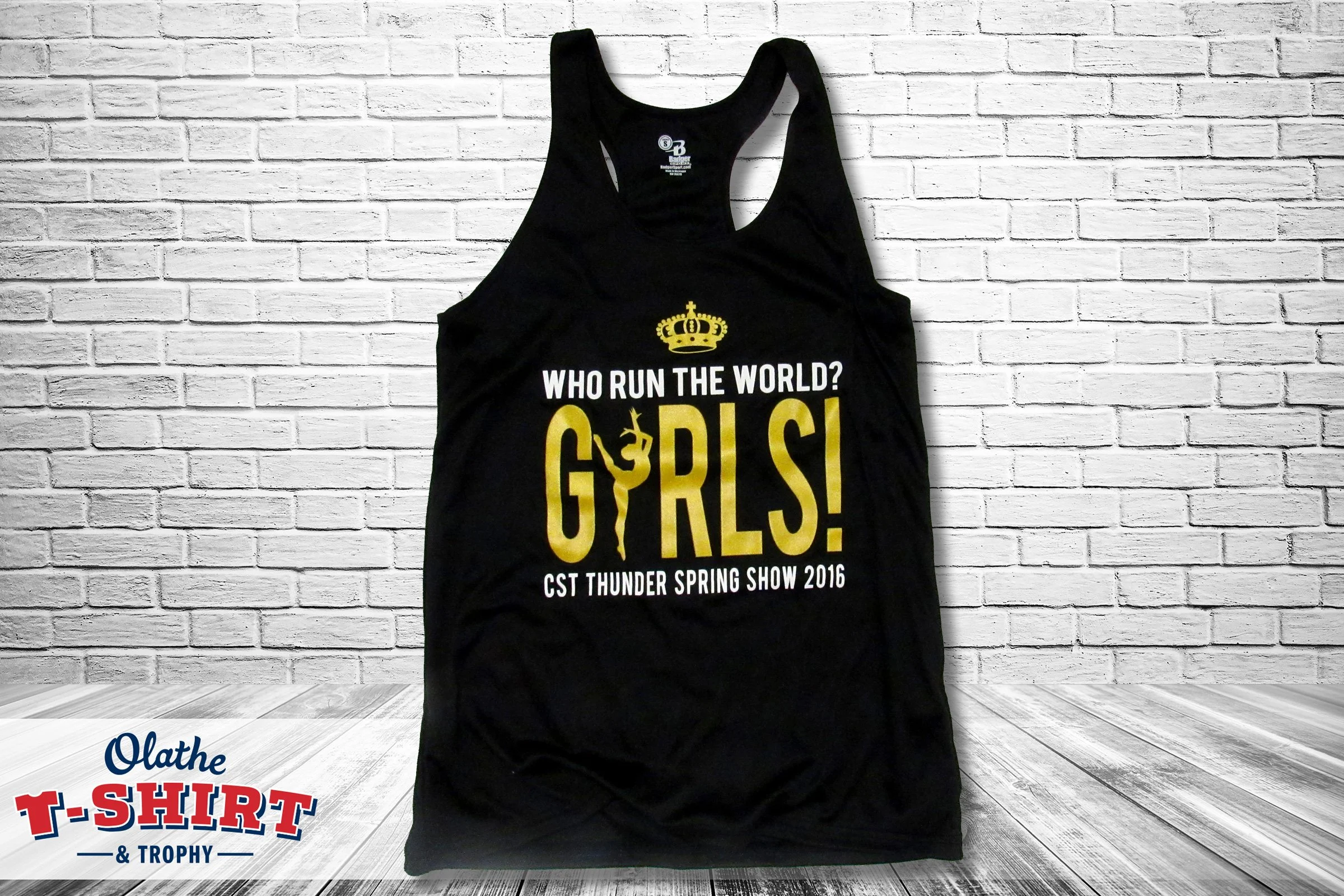 Black graphic tank top with gold and white text and a crown design, reading "Who run the world? Girls!" The tank top is hanging against a white brick wall, with a wooden floor below.