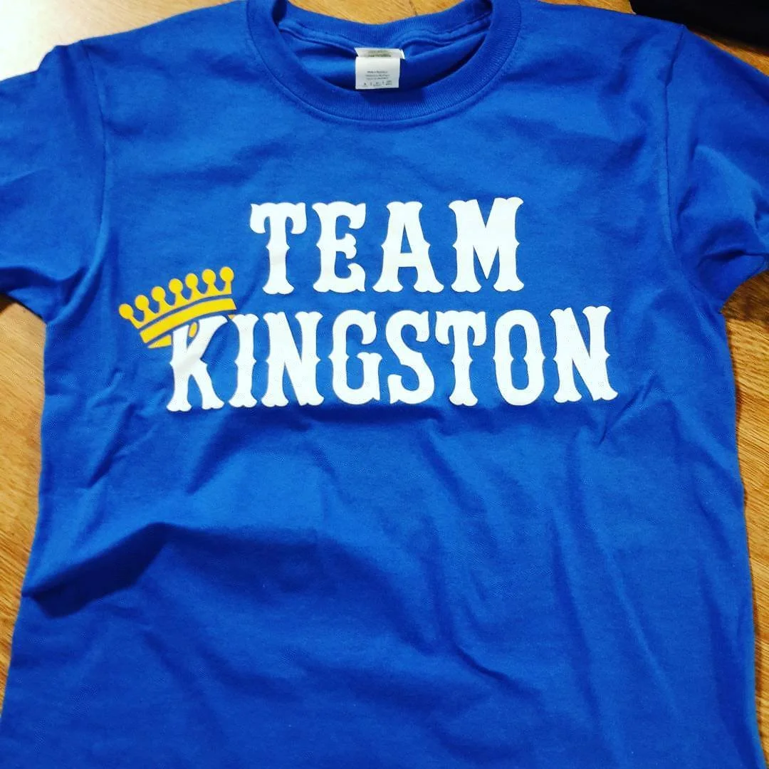 A blue T-shirt with white text that reads "TEAM KINGSTON" and a yellow crown graphic over the letter "K".