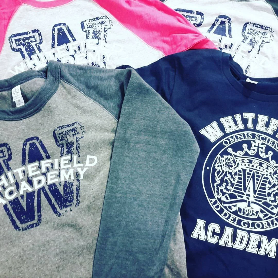 Gray and green sweatshirt, navy blue shirt, and white, pink, and gray T-shirts with 'Whitefield Academy' logos laid out on a surface.