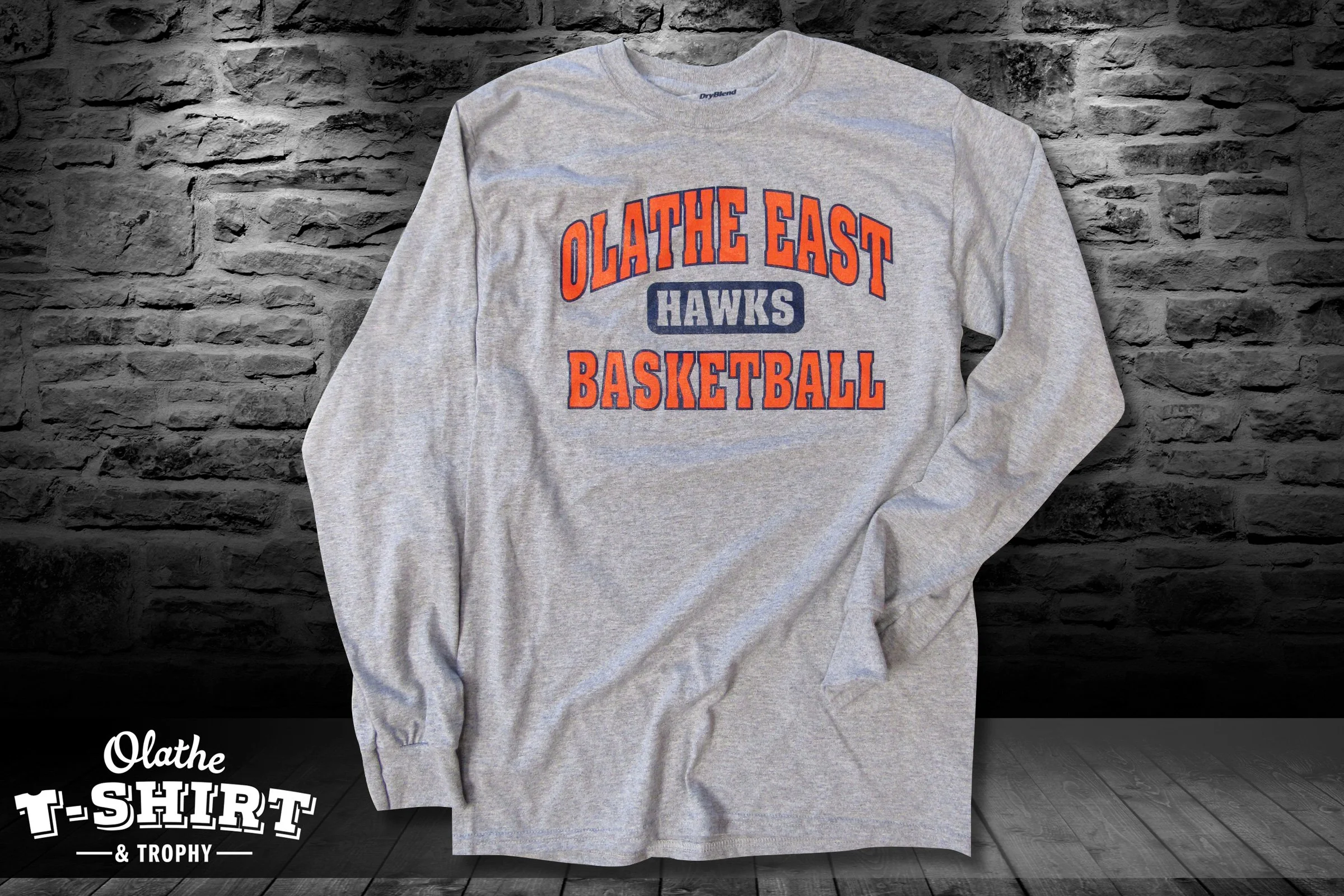 Gray sweatshirt with orange and blue text that reads "OLATHE EAST HAWKS BASKETBALL" on a dark stone wall background.