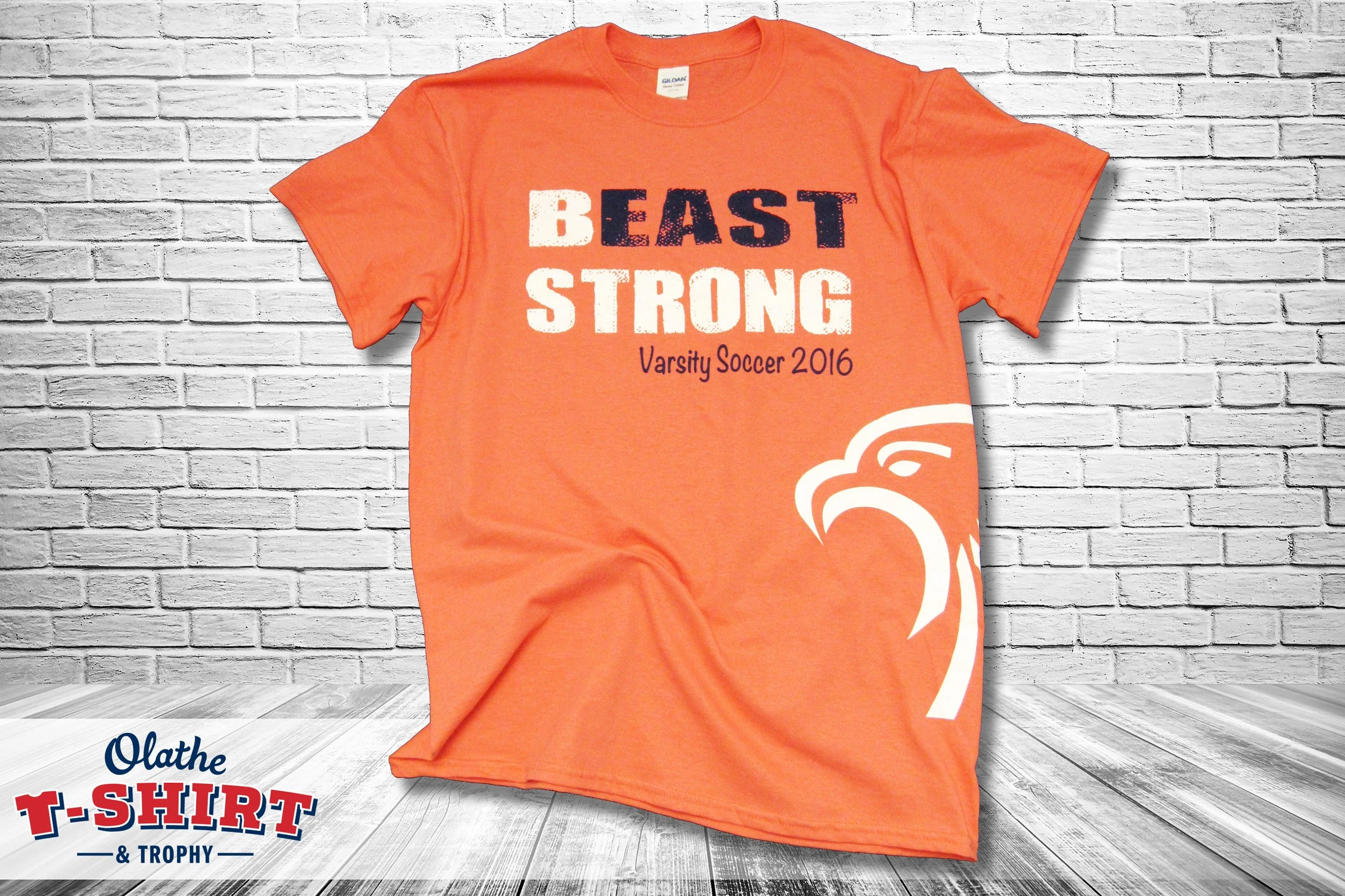 Orange T-shirt with the words 'BEAST STRONG' printed on the front, hanging against a white brick wall background.