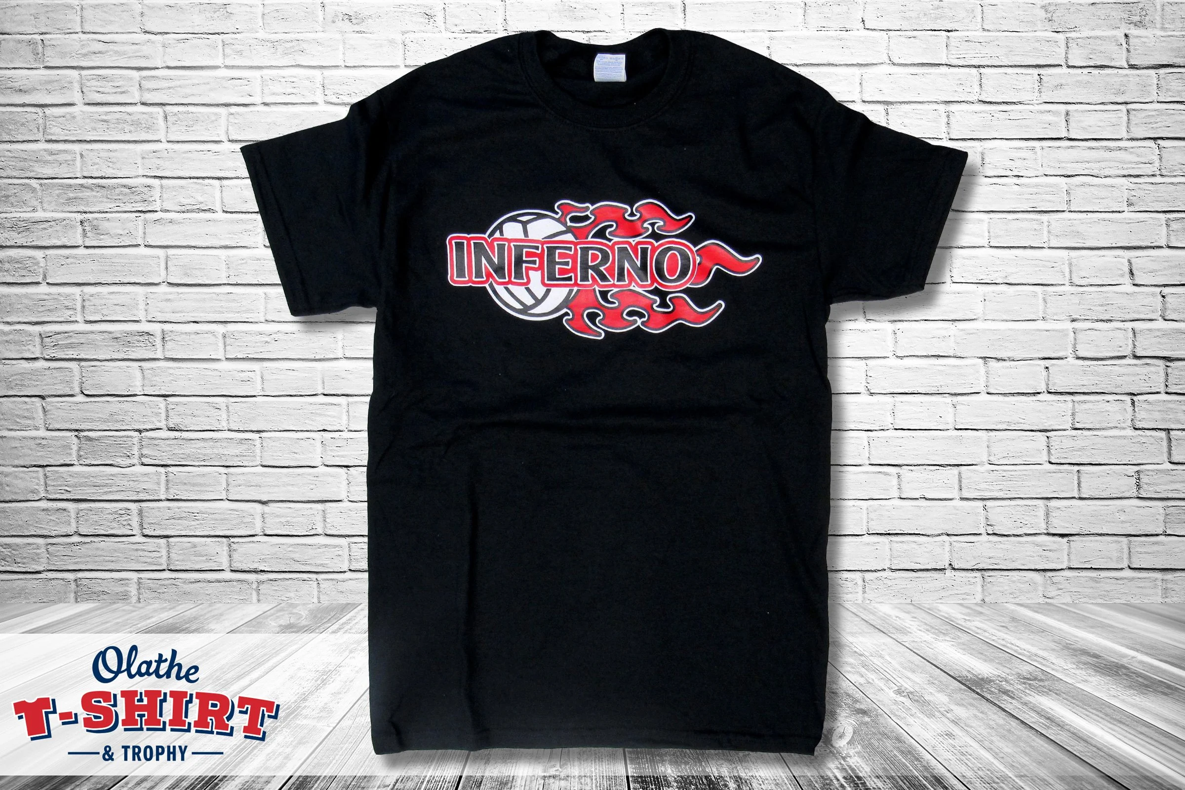 A black T-shirt with the word "INFERNO" in red and white, accompanied by a graphic of flames. The T-shirt is displayed against a white brick wall background.