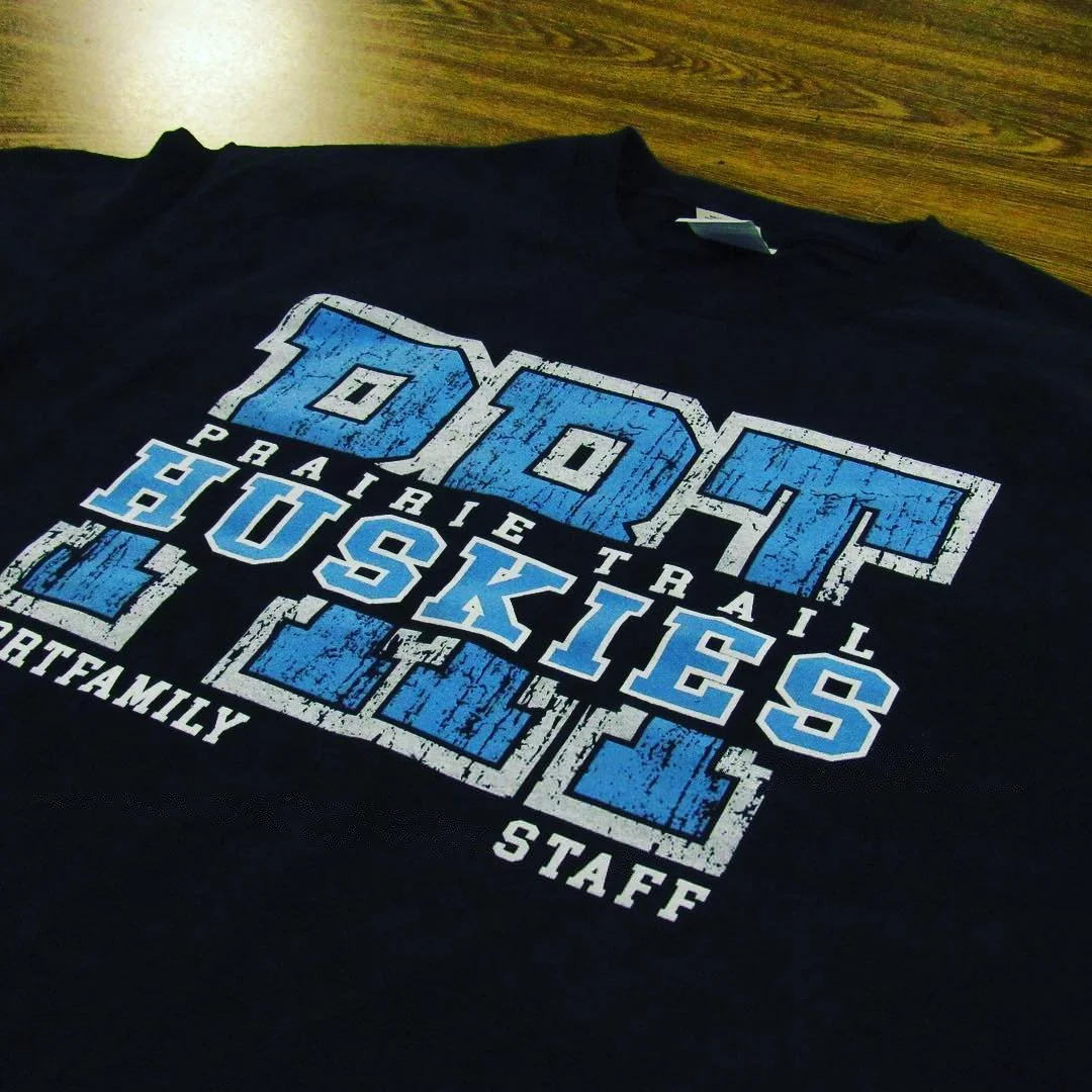 Black staff shirt with blue and white distressed school lettering.