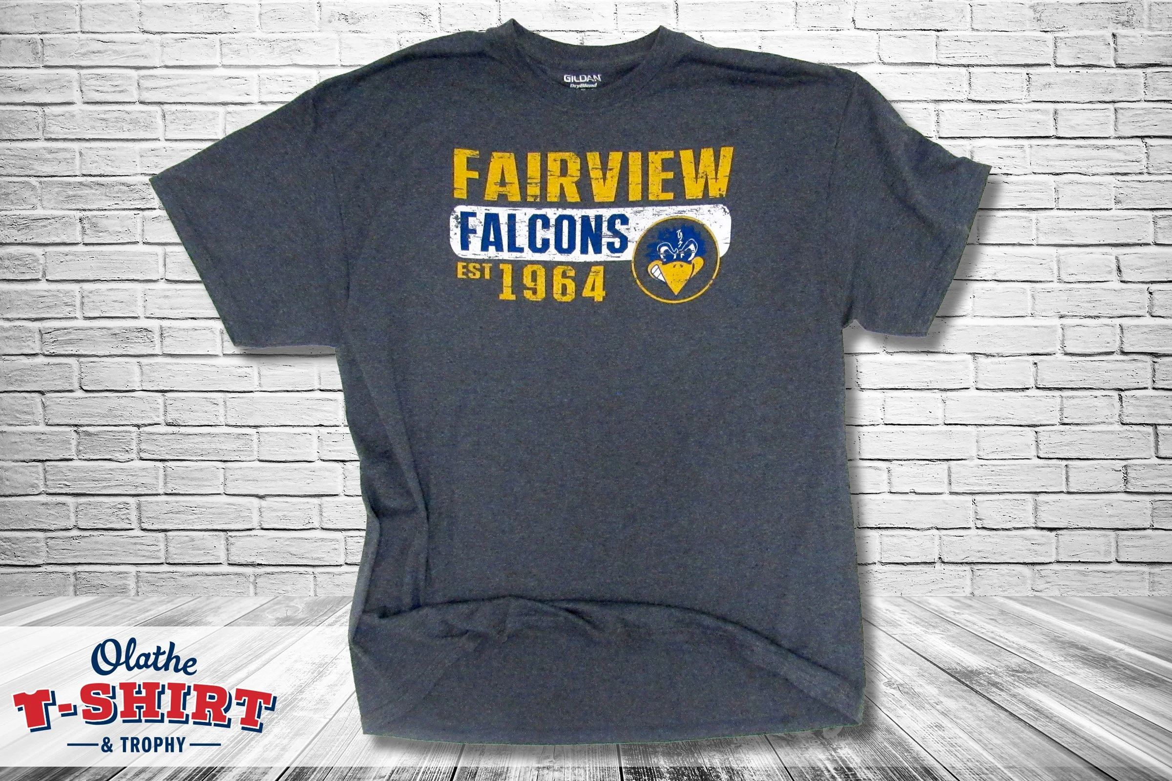 A dark gray T-shirt with yellow and blue text and a logo, hanging against a white brick wall background.