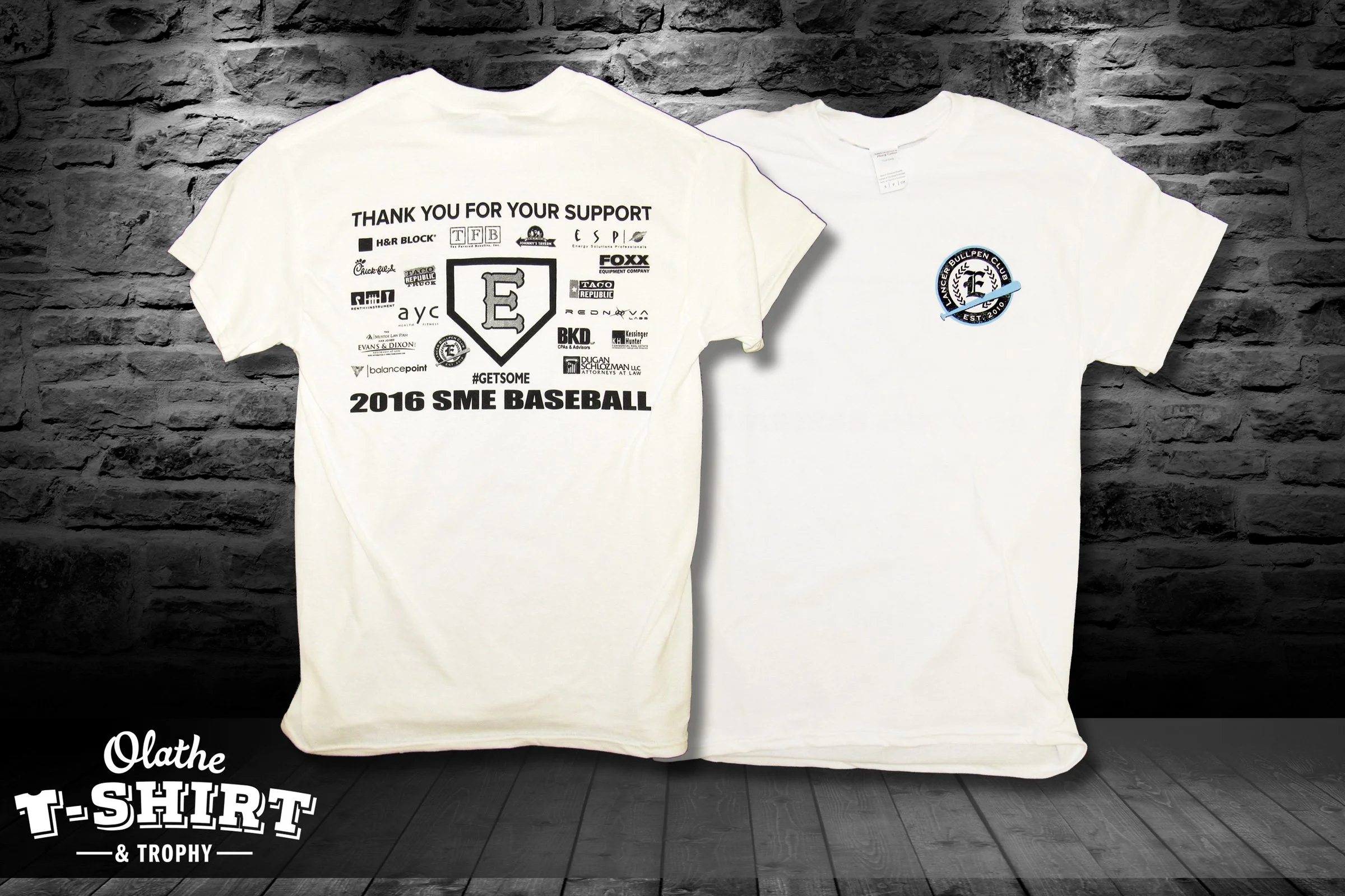 Two white T-shirts hanging on a wall with a black brick background. The shirt on the left has text and logos supporting a baseball event, and the shirt on the right has a logo of a baseball team or club. There is also some text overlay at the bottom 