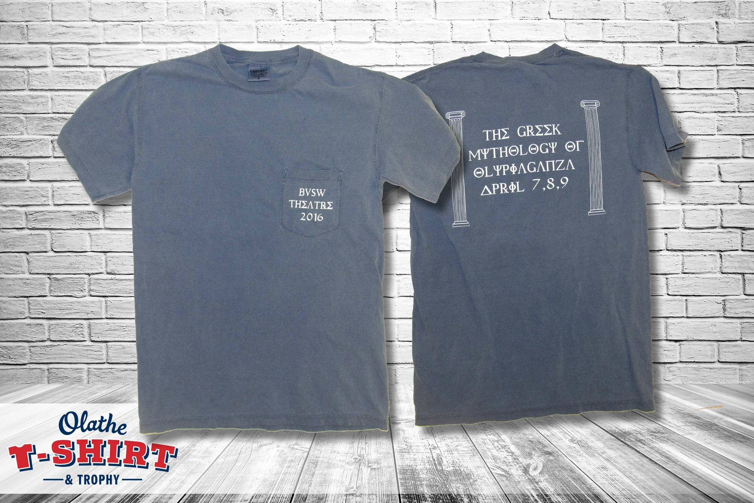 Two gray T-shirts hanging against a white brick wall. The front of one T-shirt has a small pocket with text that reads "BVSW THELTΣ 2016." The back of the other T-shirt has Greek text and images of columns with text that reads "THE GREEK MYTHOLOGY OF
