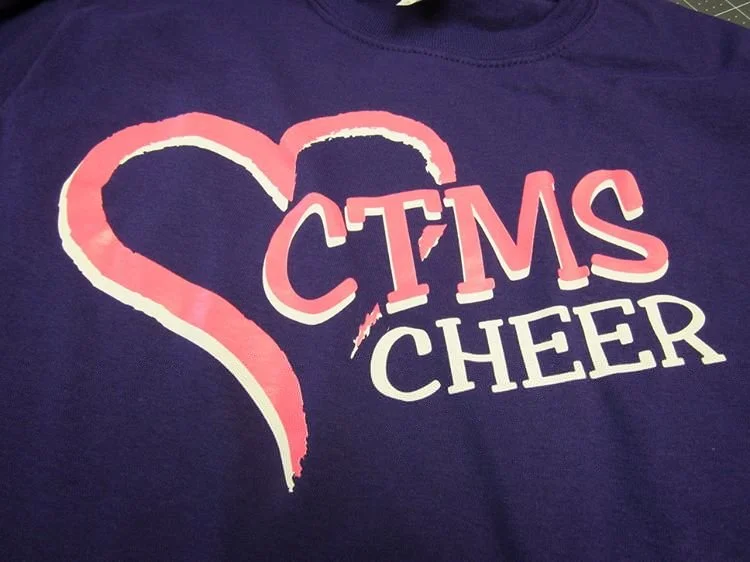 A purple T-shirt with pink and white text that reads 'CTMS Cheer' and a large pink heart outline.
