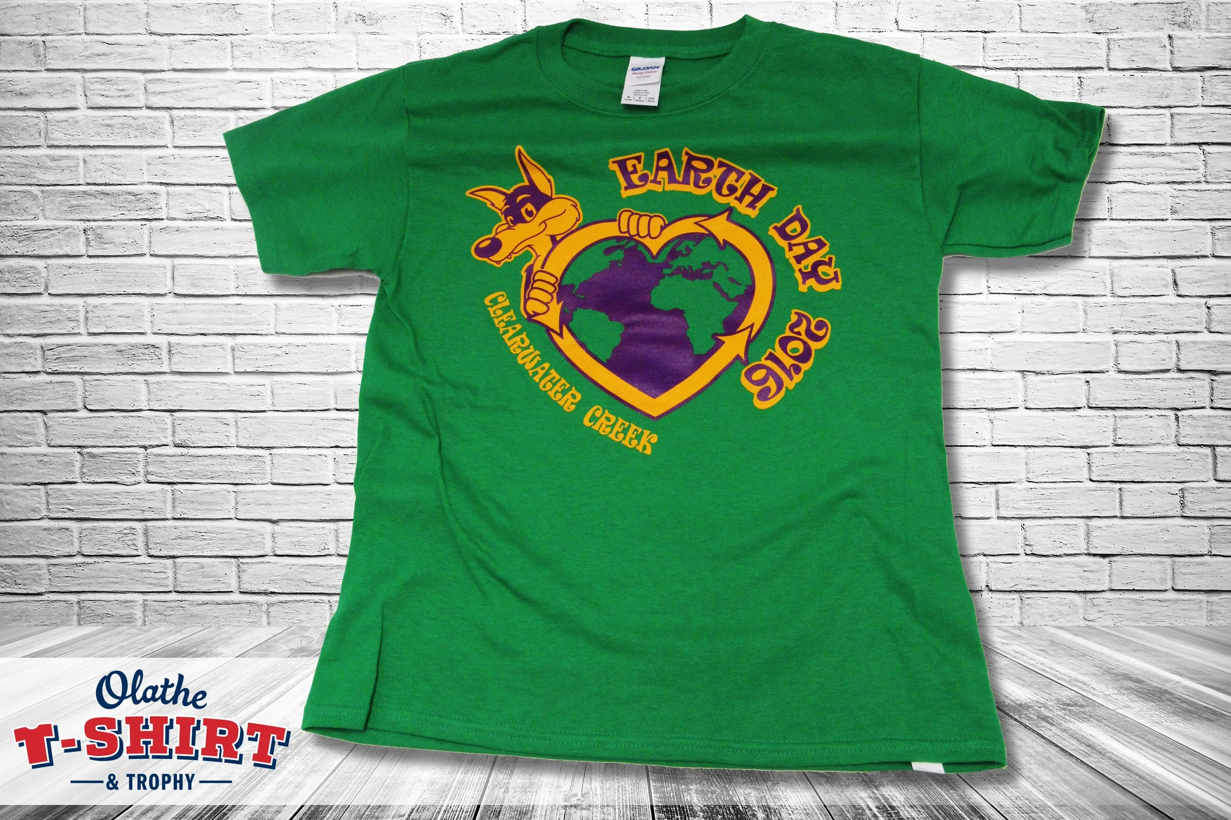 Green T-shirt featuring a cartoon coyote holding a heart-shaped world map, with yellow and purple text reading 'Earth Day' and 'Clevelandier Creek 2016'.