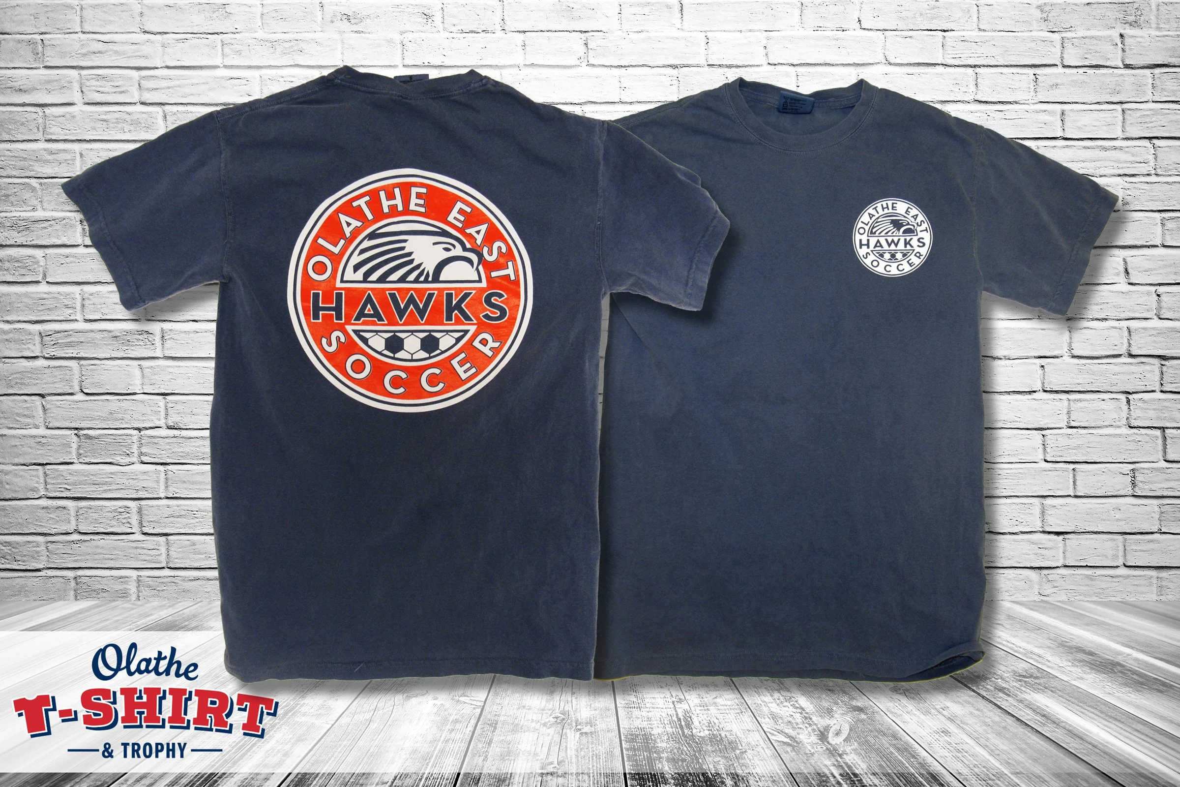 Two navy blue T-shirts hanging against a white brick wall. The front of the shirts bear the logo of Olathe East Hawks Soccer, with one prominently displaying the logo and the other showing a smaller version. The background has a white brick texture, 