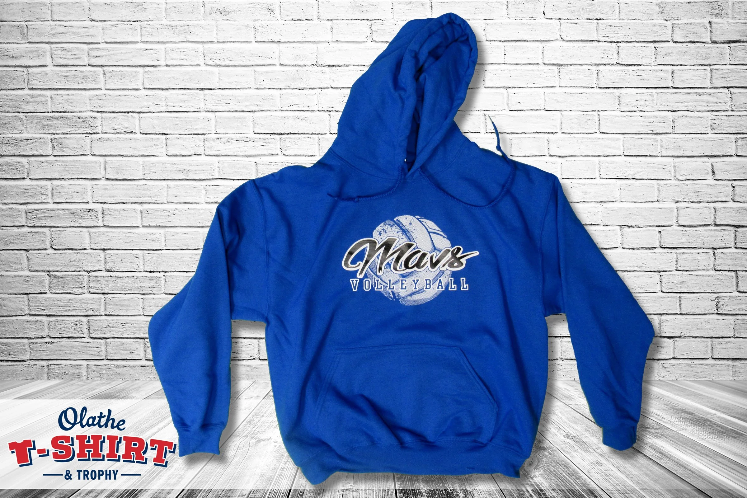 A blue volleyball hoodie with the word 'Mavs' and a volleyball graphic on the front, hanging against a white brick wall.