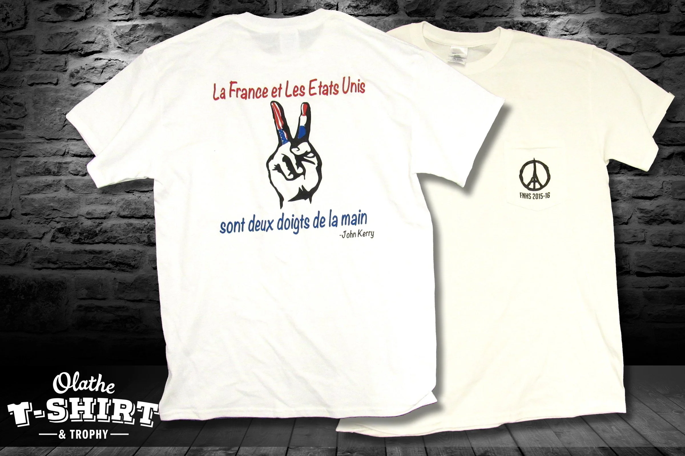 Two white T-shirts with printed designs are hanging against a brick wall. The shirt in front features a hand making a peace sign with a French and American flag motif, surrounded by French and American text. The second shirt has a peace symbol with t