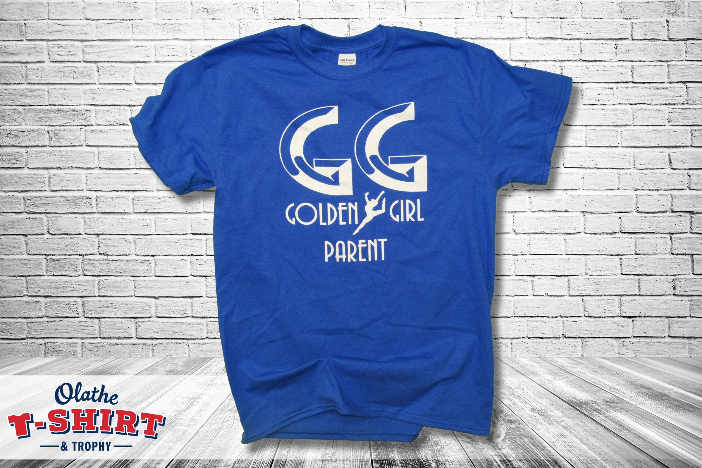 A blue T-shirt hanging against a white brick wall, with a logo featuring two mirrored G's and the words 'Golden Girl Parent' printed on it.