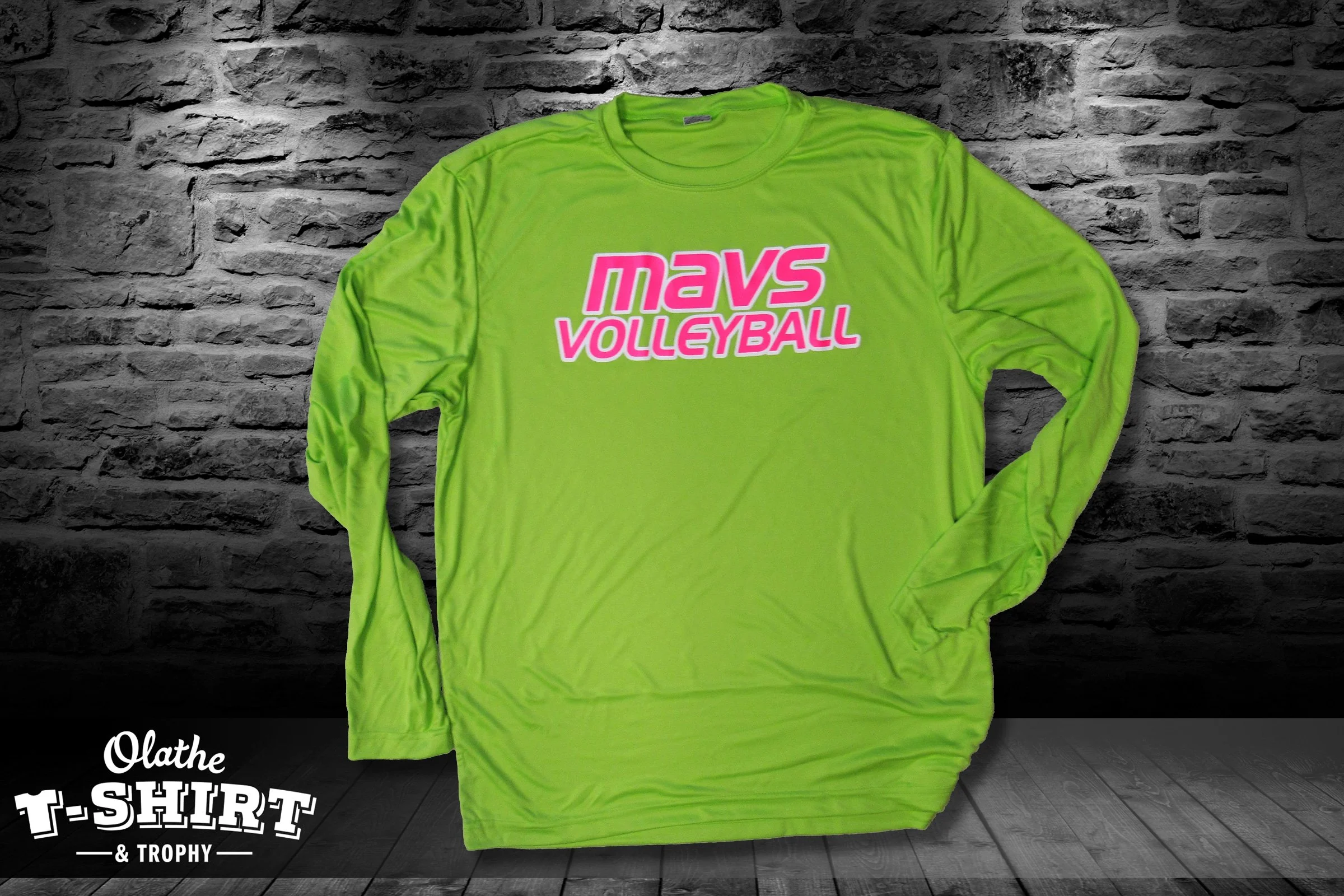 Bright lime green long-sleeve volleyball T-shirt hanging on a dark wooden floor and against a gray brick wall.