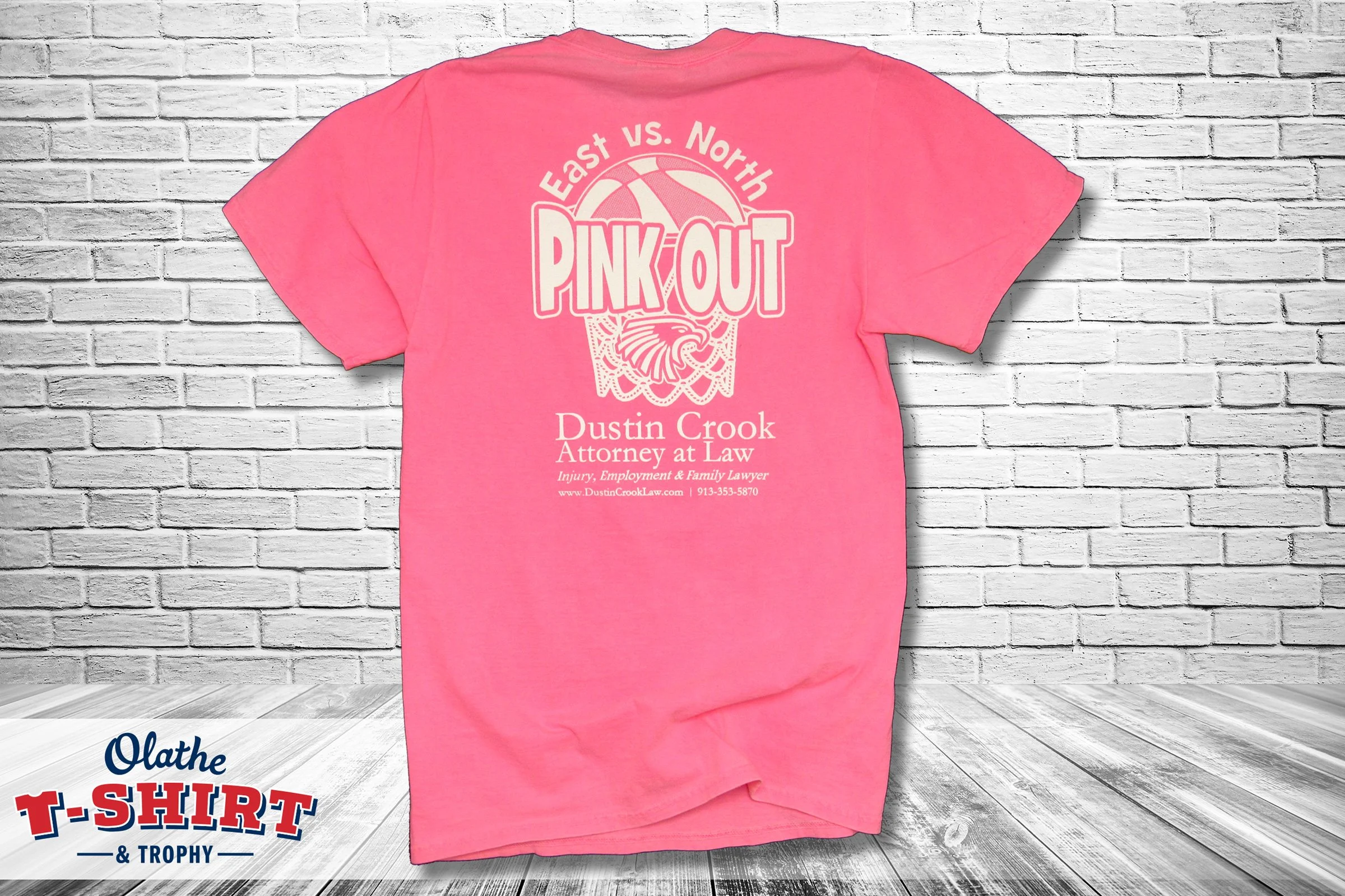 Pink T-shirt hanging against a white brick wall with a basketball graphic and the words "East vs. North Pink Out" printed on the back.
