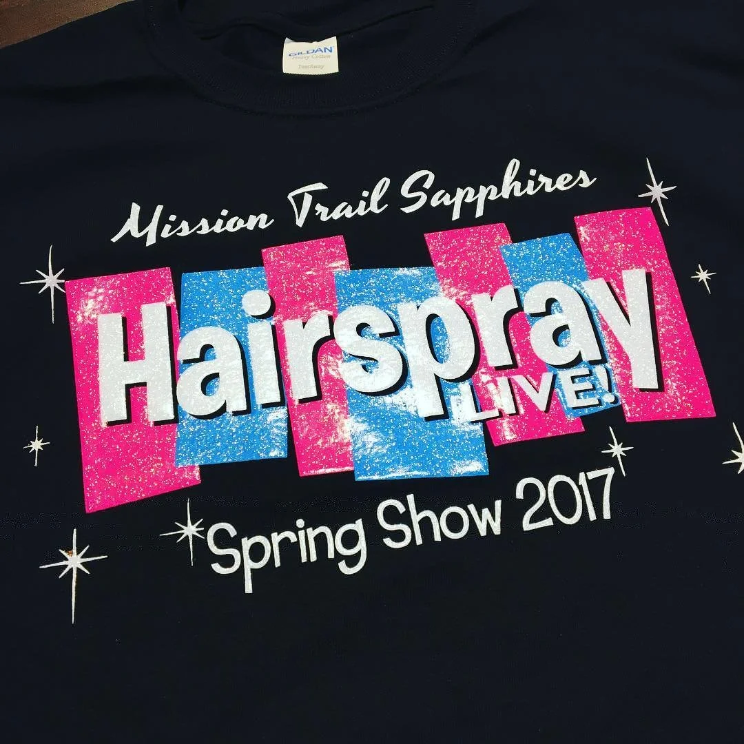 Pink and blue graphic design on a black shirt promoting Hairspray Live! Spring Show 2017 by Mission Trail Sapphires.