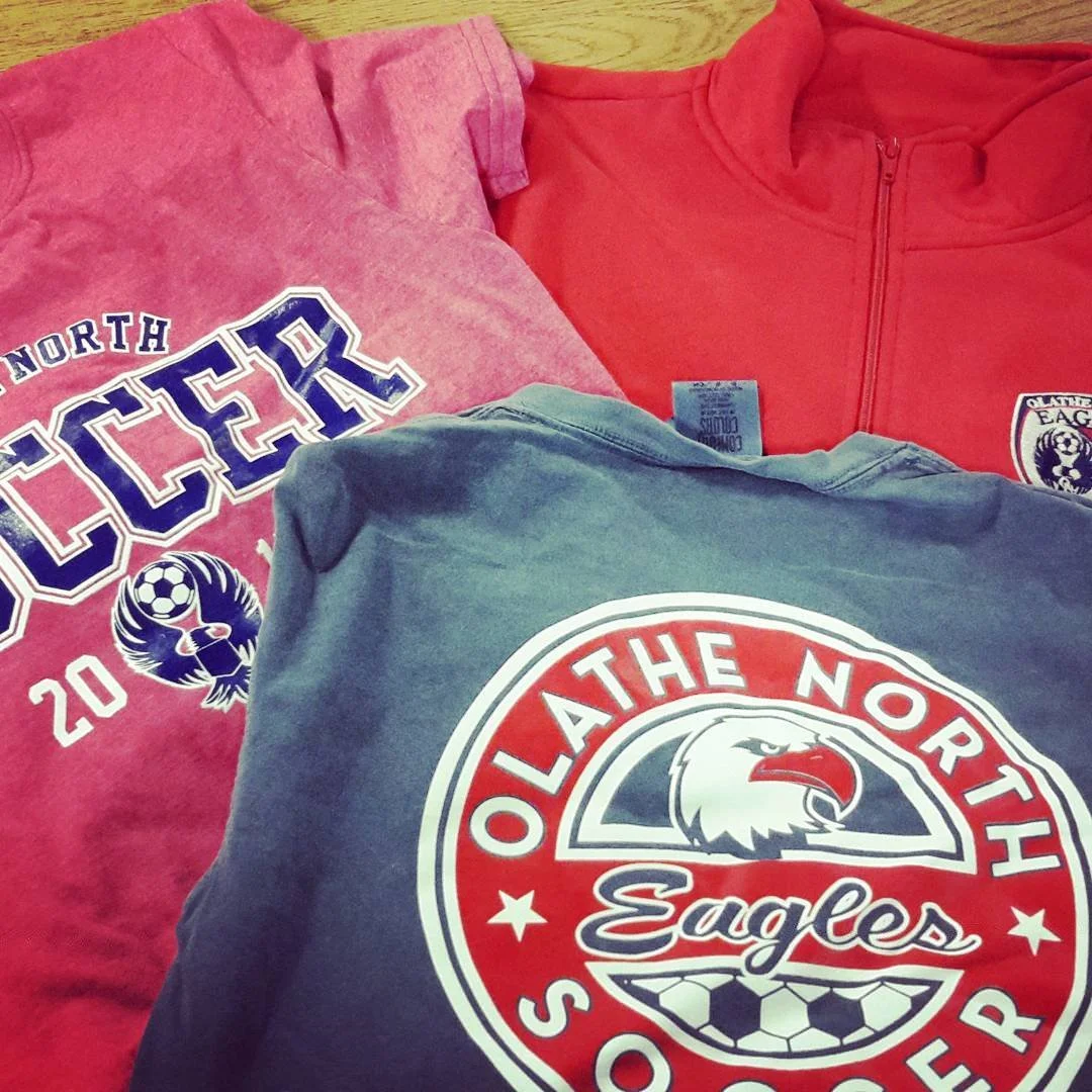 Three folded sweatshirts with sports team logos and names, one red, one gray, and one pink, on a wooden surface.