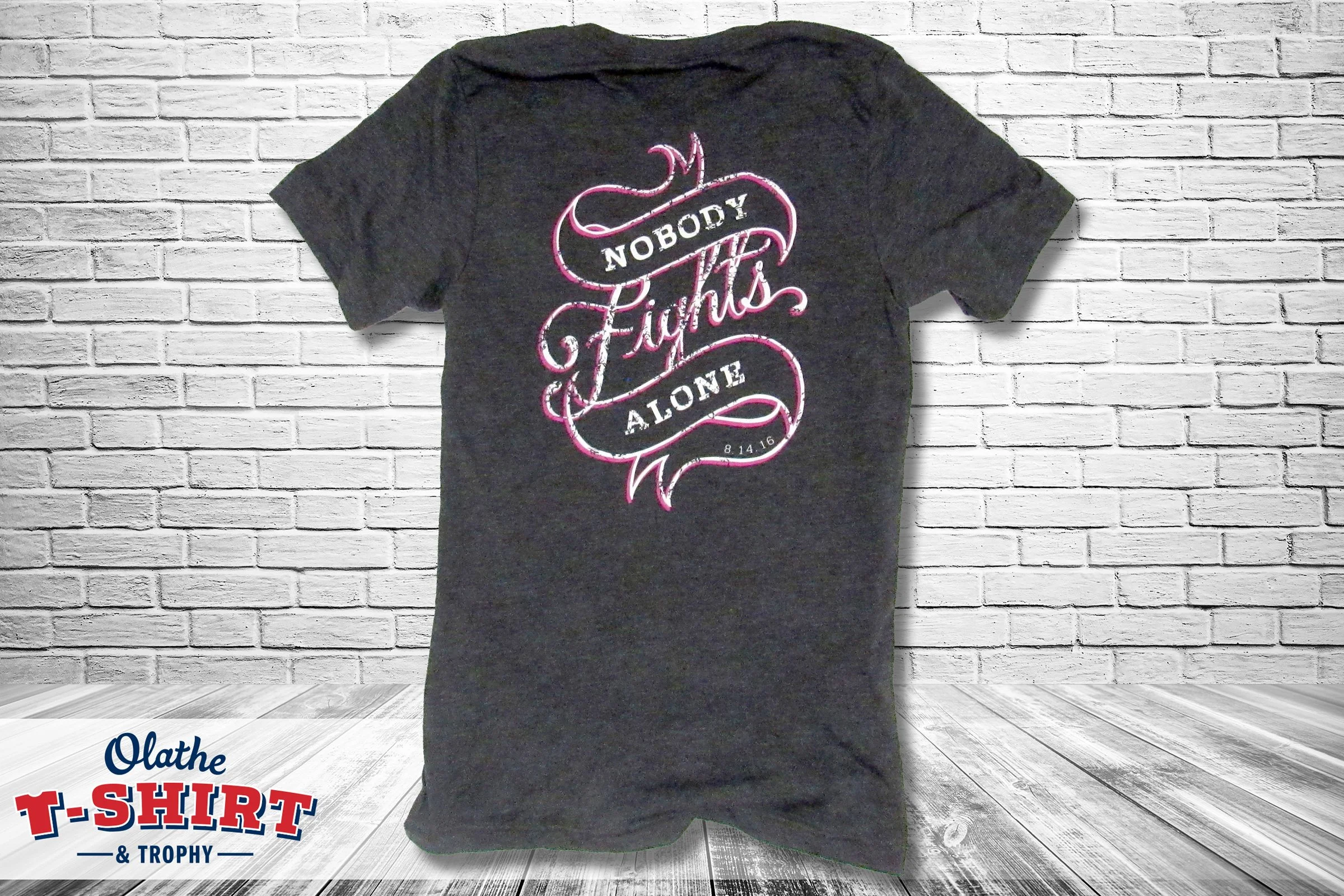 Black Nobody Fights Alone shirt with pink and white lettering, displayed against a white brick wall.