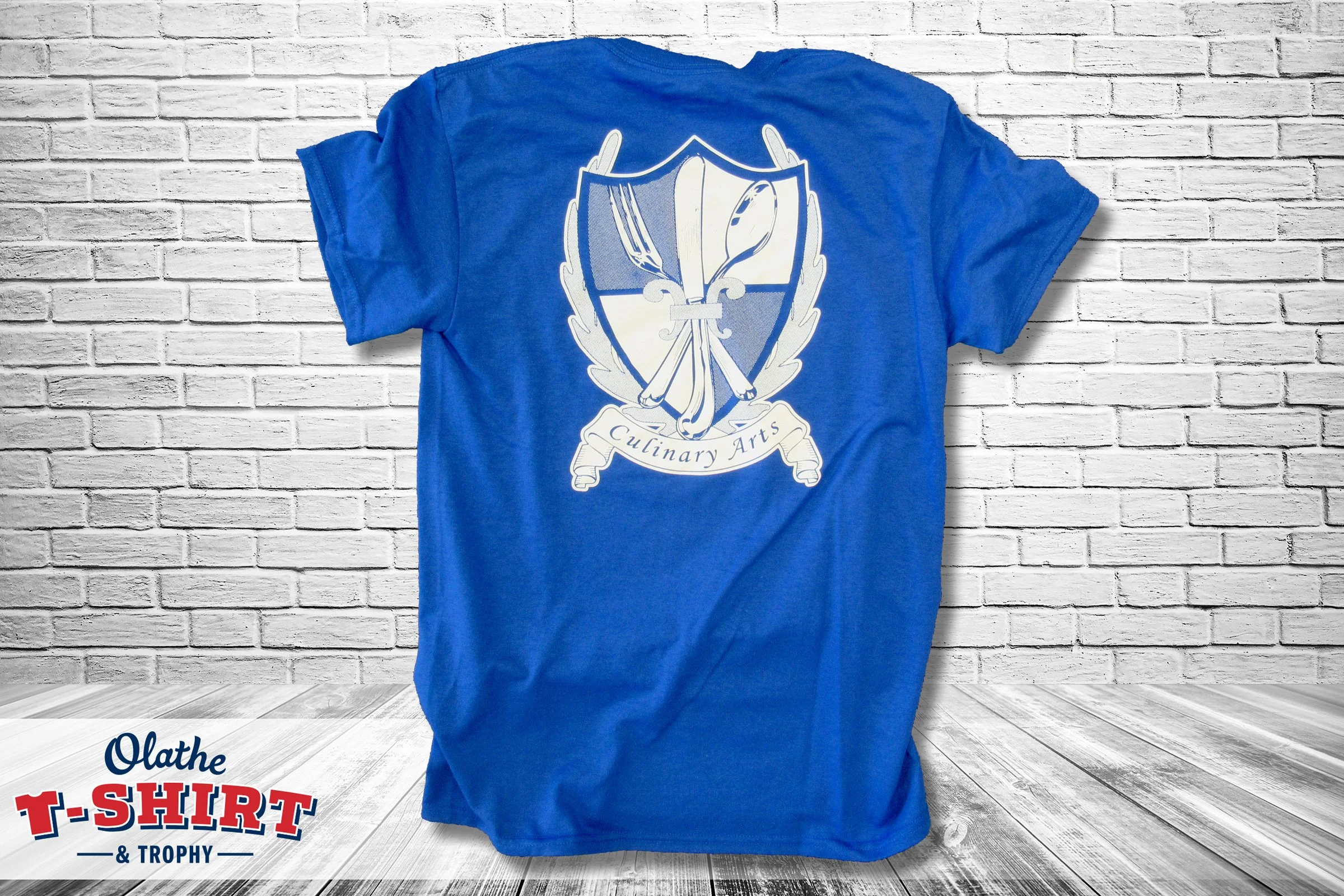 Blue T-shirt hanging against a white brick wall with a crest-like emblem featuring a feather, a spoon, a fork, and a fleur-de-lis, and the words 'Culinary Arts' underneath.