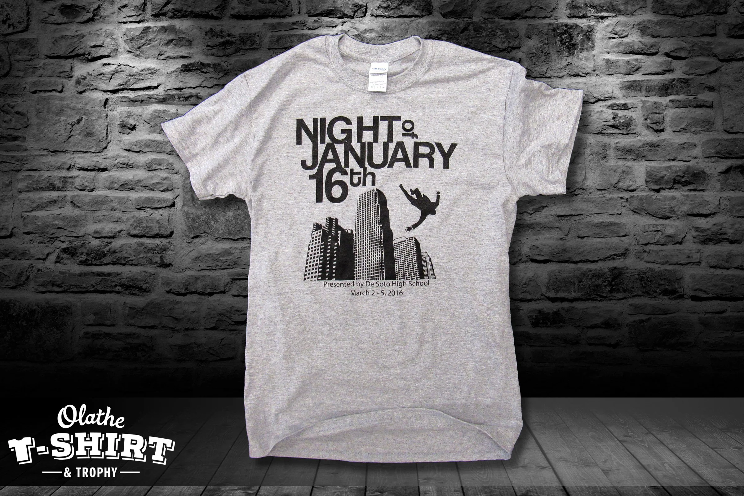 Gray T-shirt with black text and graphics, attached to a gray background. The text reads "Night of January 16th" with additional details below. The graphic shows a city skyline and a falling person.