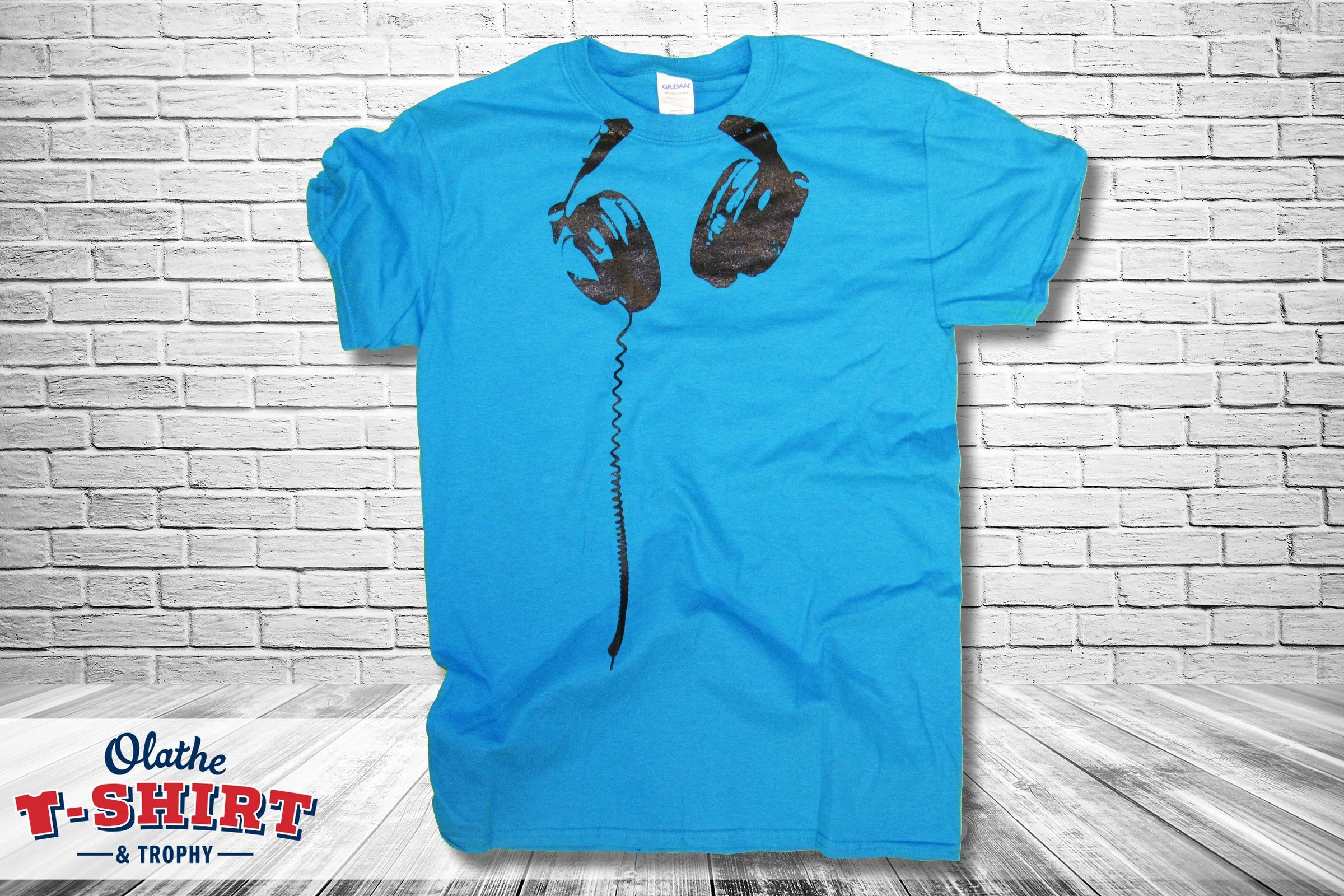Blue T-shirt with a graphic of two black headphones on the front, hanging against a white brick wall background.
