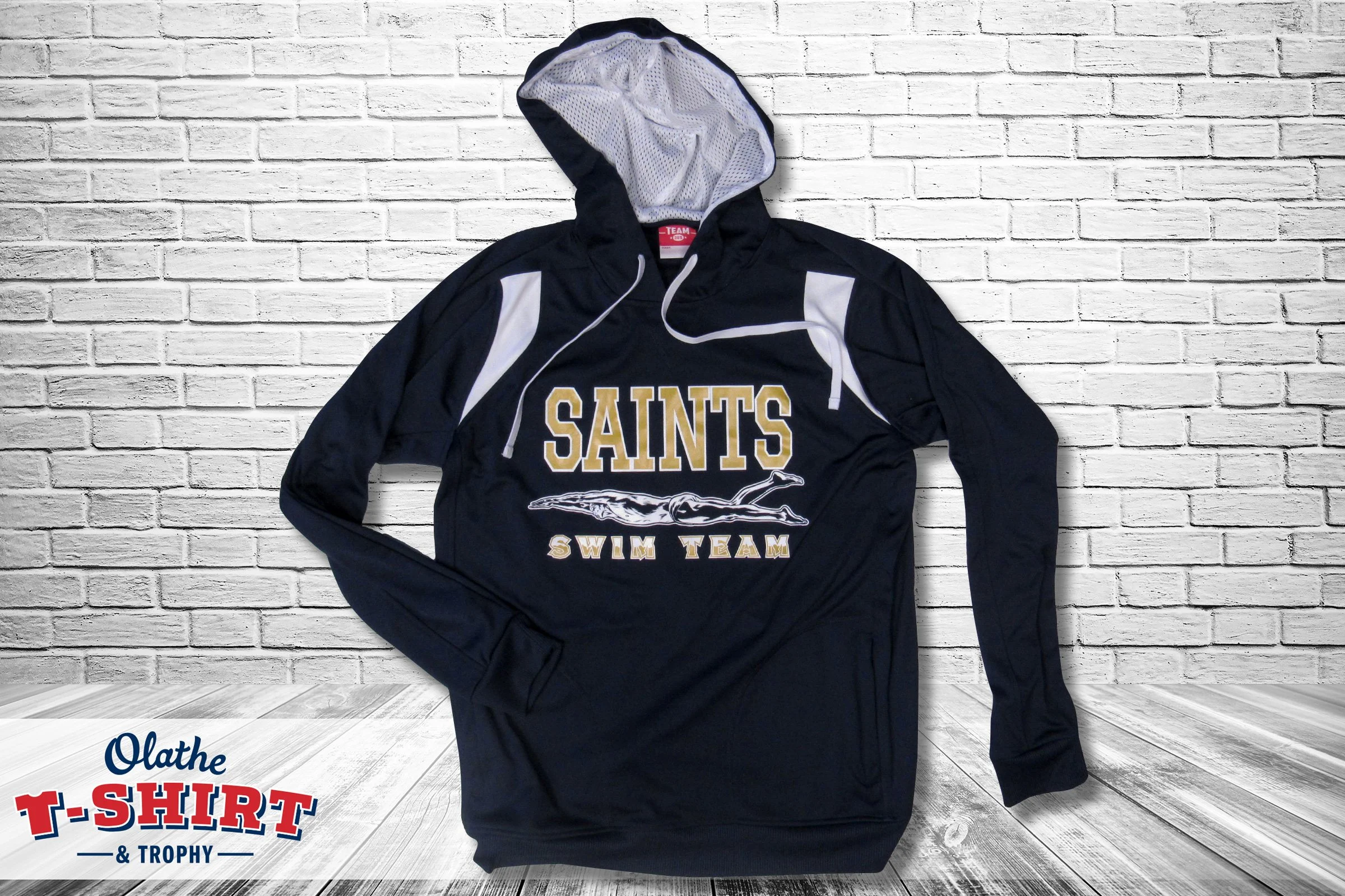 A navy blue hoodie with white accents and the text 'SAINTS SWIM TEAM' on the front, lying on a white brick wall background.