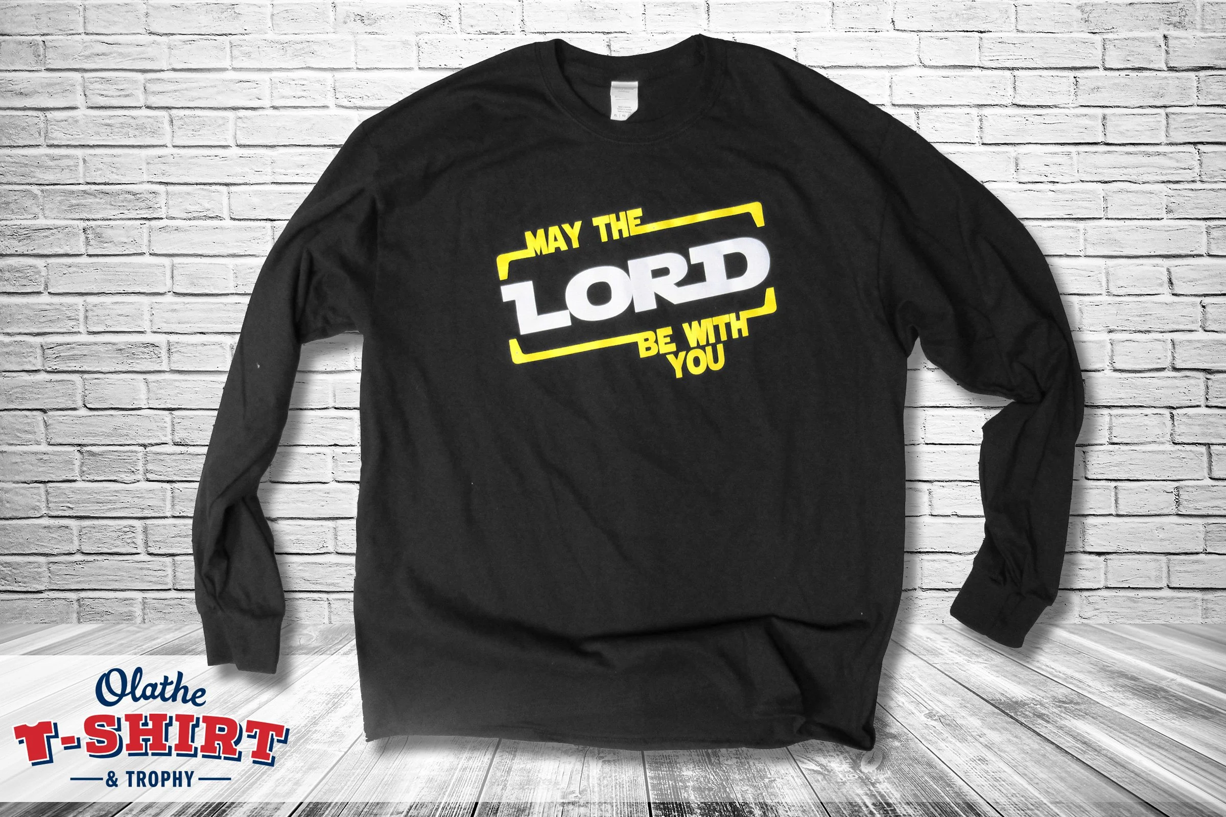 A black long-sleeve T-shirt with yellow and white text that says "May the LORD be with you," displayed against a white brick wall background.