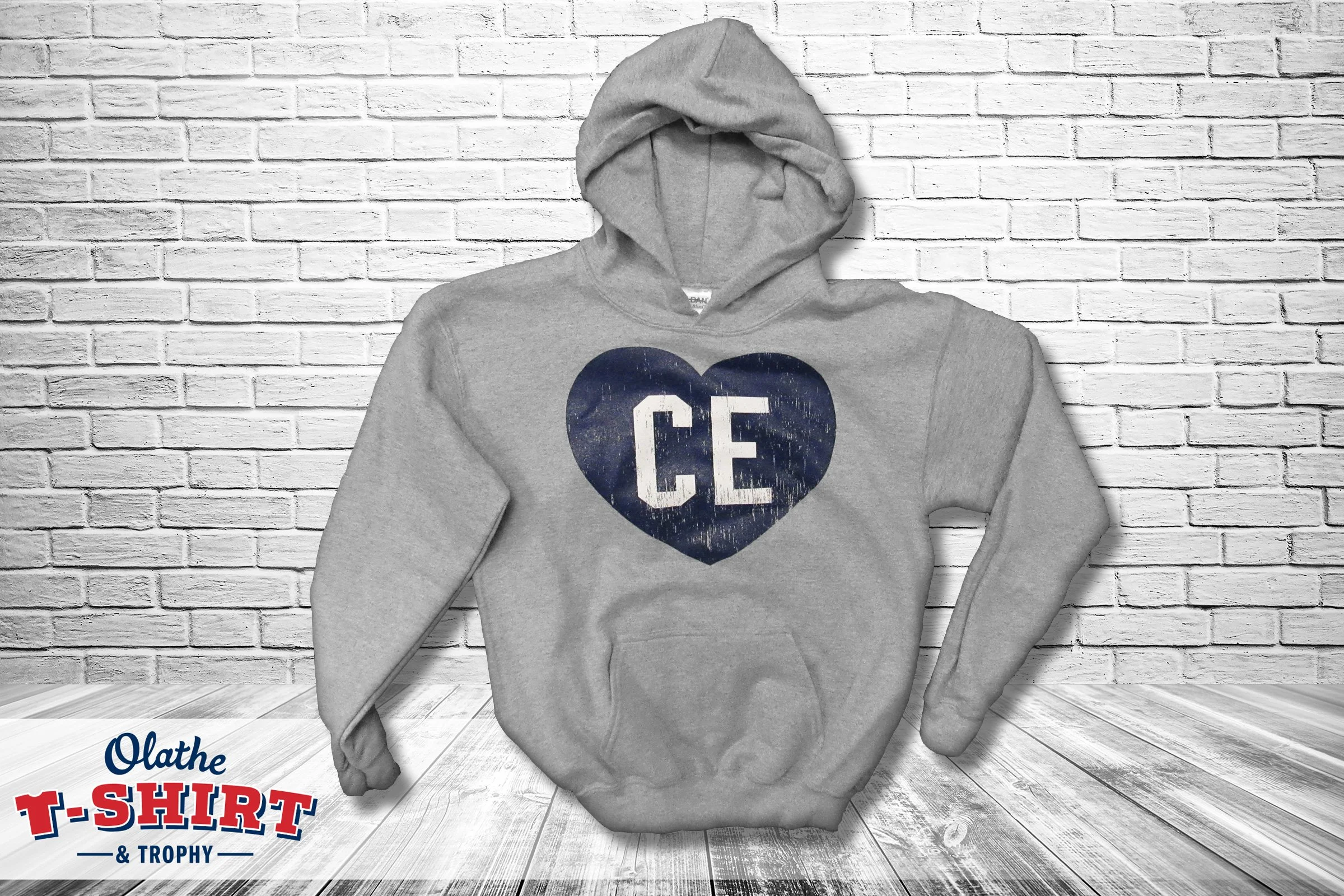 Gray hoodie sweatshirts hanging on a white brick wall background with text in the bottom corner that reads 'Olathe T-Shirt & Trophy'.