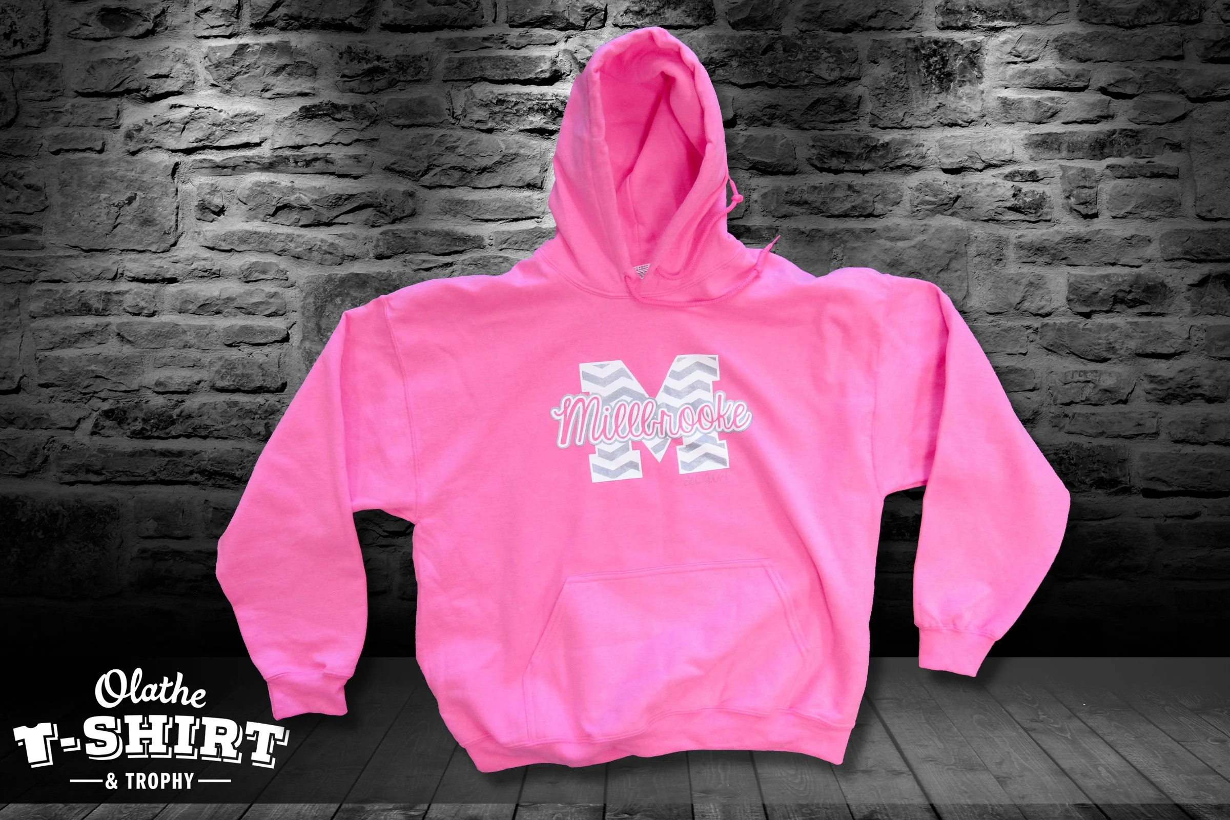 Pink hoodie with a chevron patterned letter 'M' and the name 'Millbrooke' printed on it, hanging against a gray brick wall background.