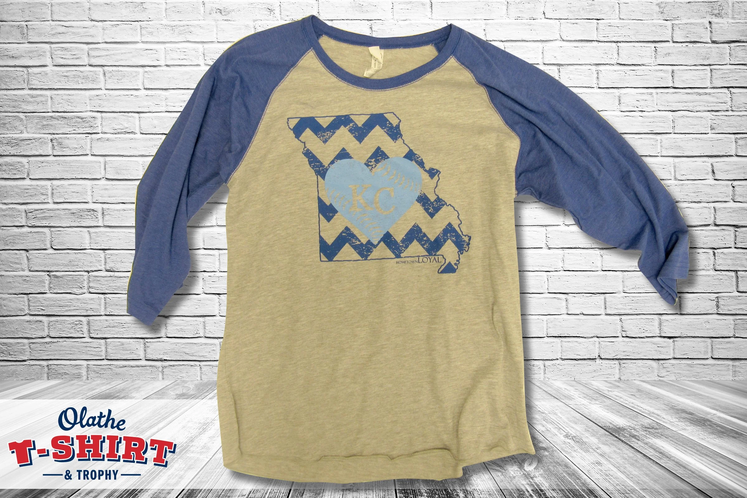 A baseball-style long-sleeve shirt with blue sleeves and a light beige body, featuring a blue heart with KC inside and a map outline of Kansas in the background. The shirt is hanging against a white brick wall background.