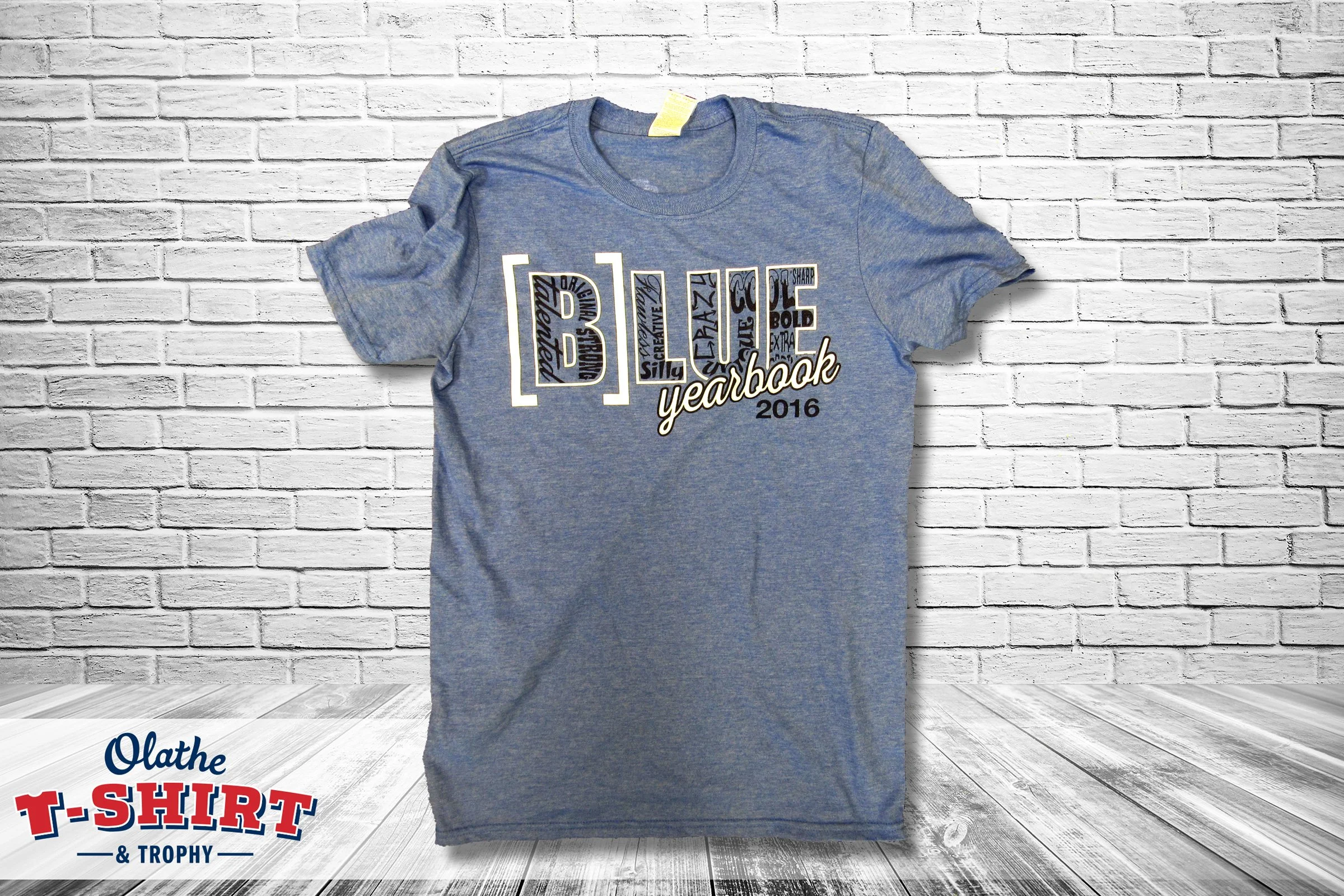 A blue t-shirt hanging on a wall with white brick background, containing decorative text and graphics for a 2016 yearbook.