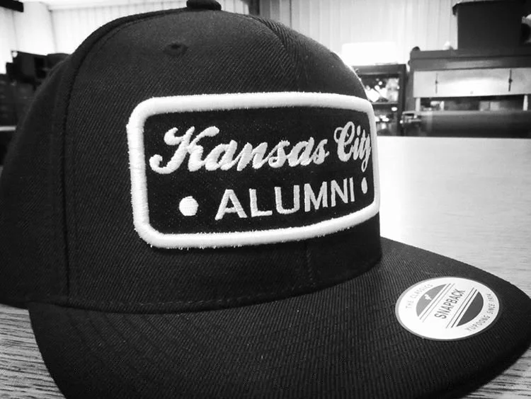 A black baseball cap with a patch that reads "Kansas City Alumni" on the front. The cap is on a wooden table with some furniture in the background.