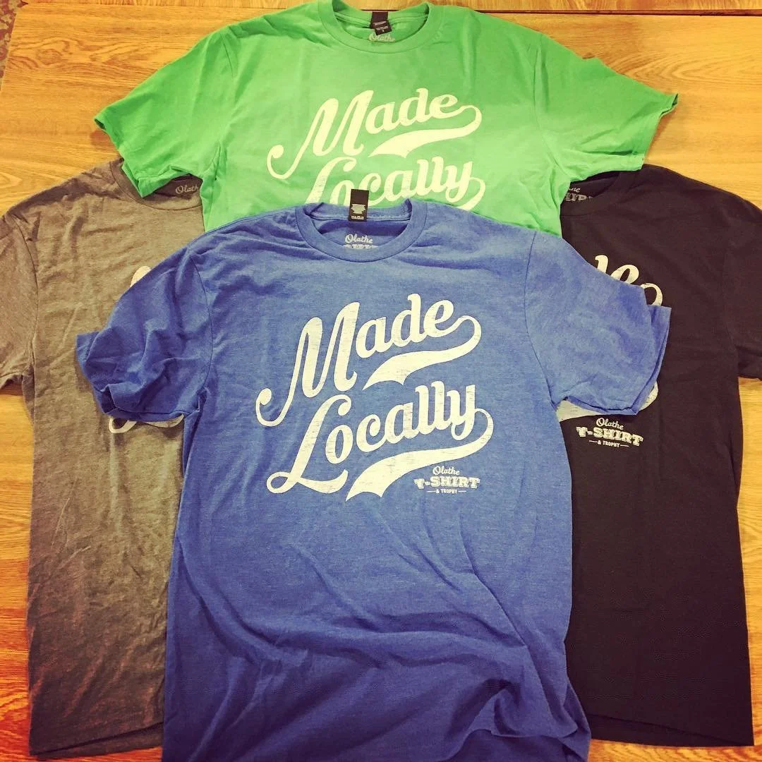 Four colored T-shirts (green, blue, gray, black) with the phrase 'Made Locally' printed on the front, displayed on a wooden surface.