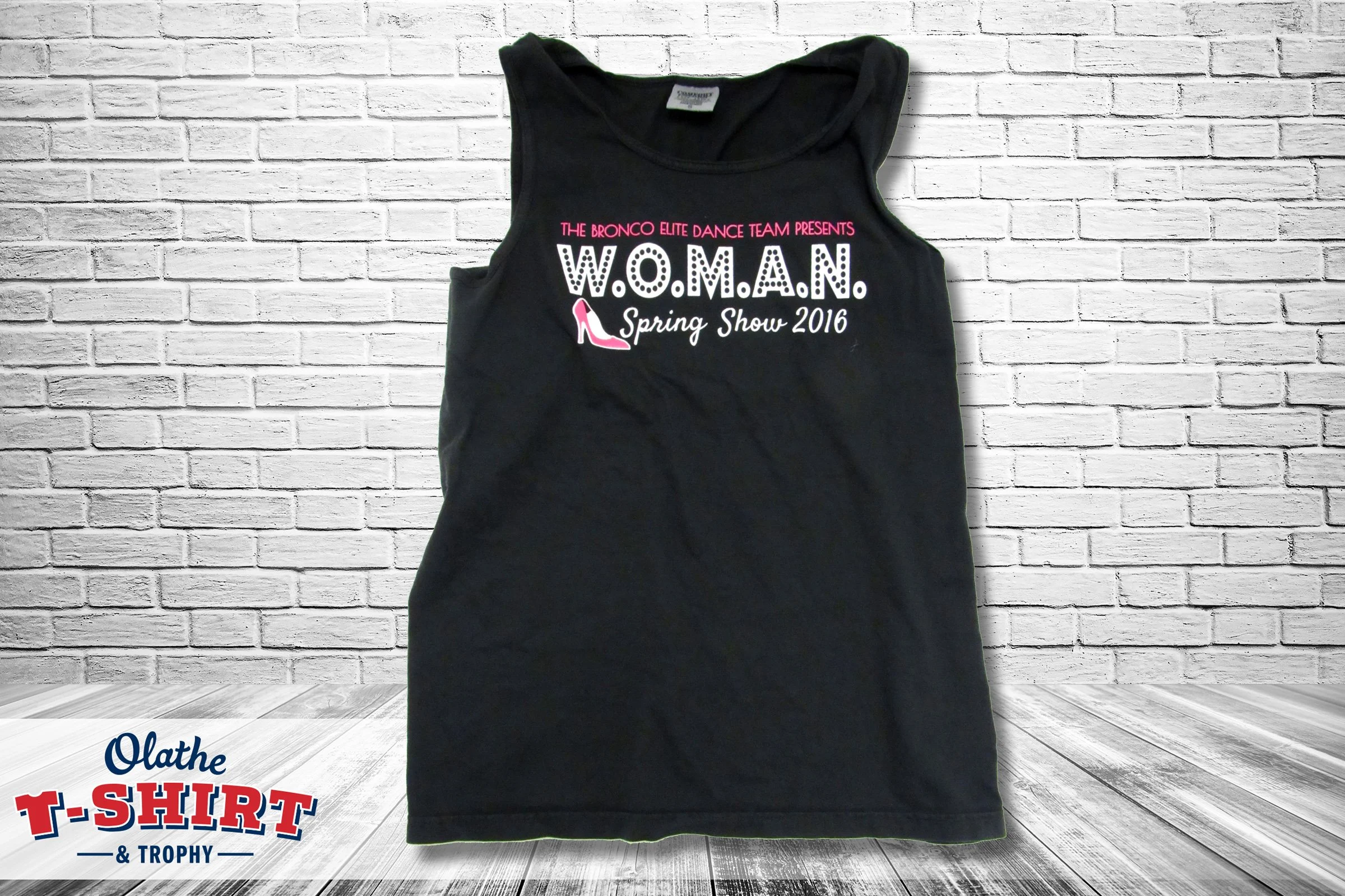 Black sleeveless T-shirt with pink, white, and black printed text, displayed against a white brick wall background.