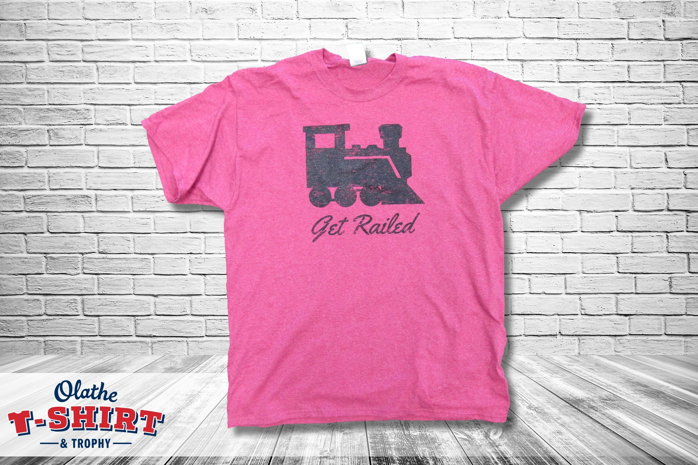 Pink T-shirt with a silhouette of a train and the words "Get Railed" printed on it, hanging in front of a white brick wall.