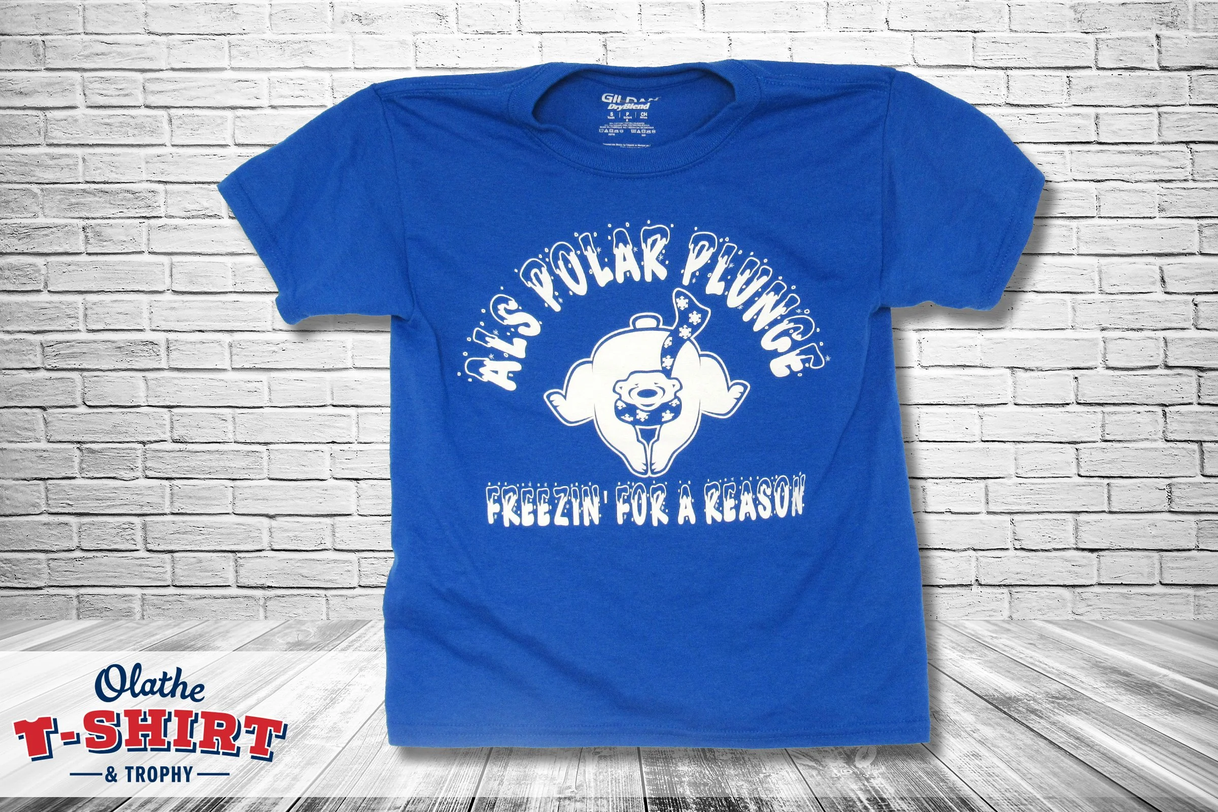A blue T-shirt with the text 'A Little Polar Plunge' and an illustration of a cartoon polar bear wearing a Santa hat and scarf, against a white brick wall background.