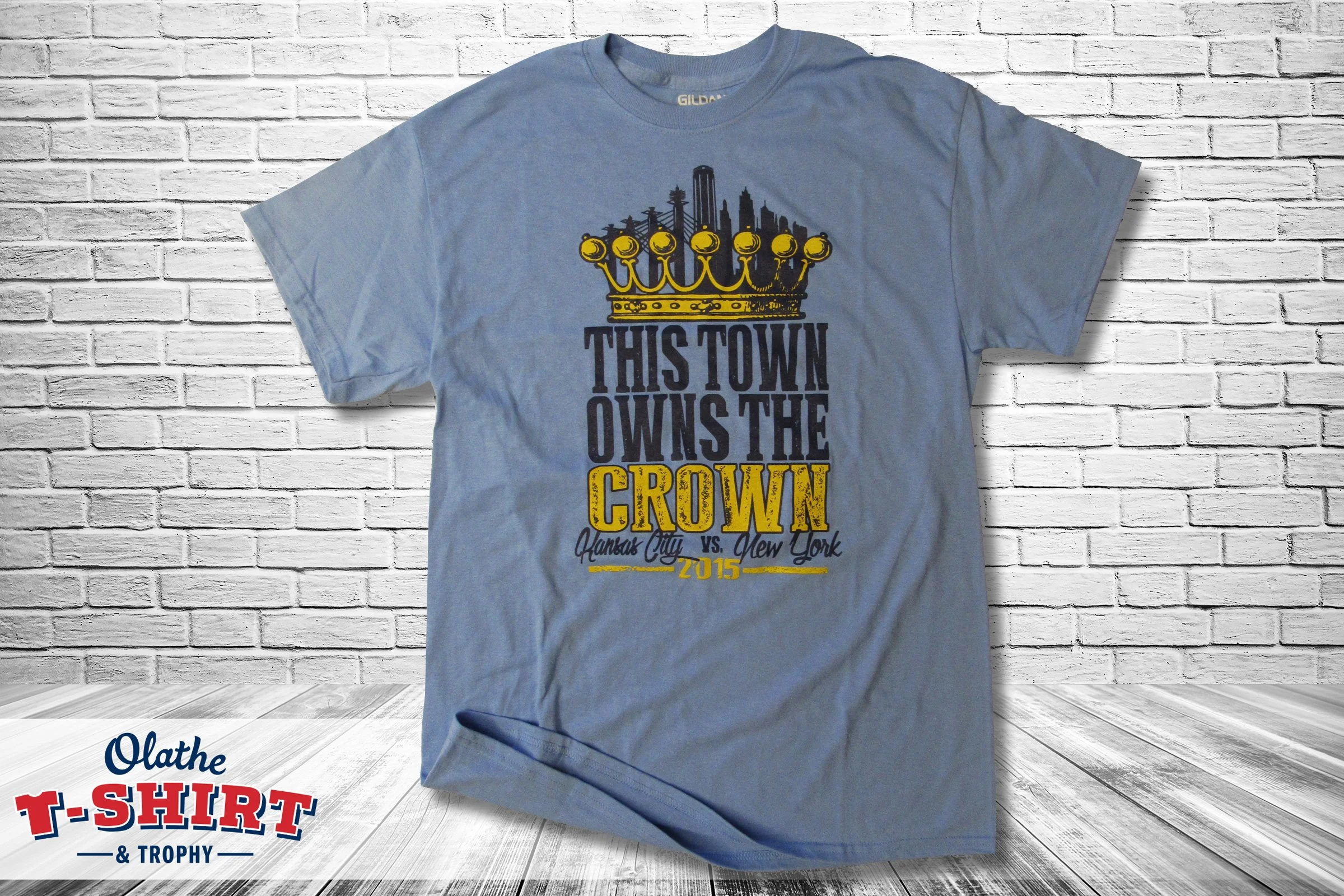 Light blue T-shirt with a yellow crown graphic and black text that reads, "This town owns the crown, Kansas City vs. New York 2015," hung on a white brick wall.