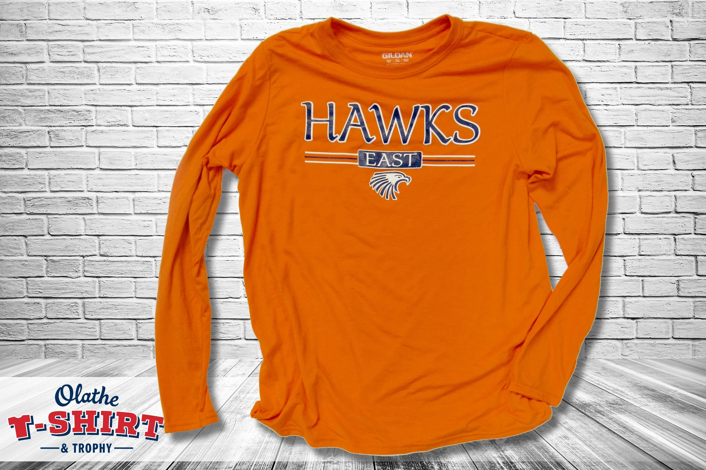 An orange long-sleeve T-shirt with the words "HAWKS EAST" and a hawk logo printed on it, laid out on a white brick wall background.