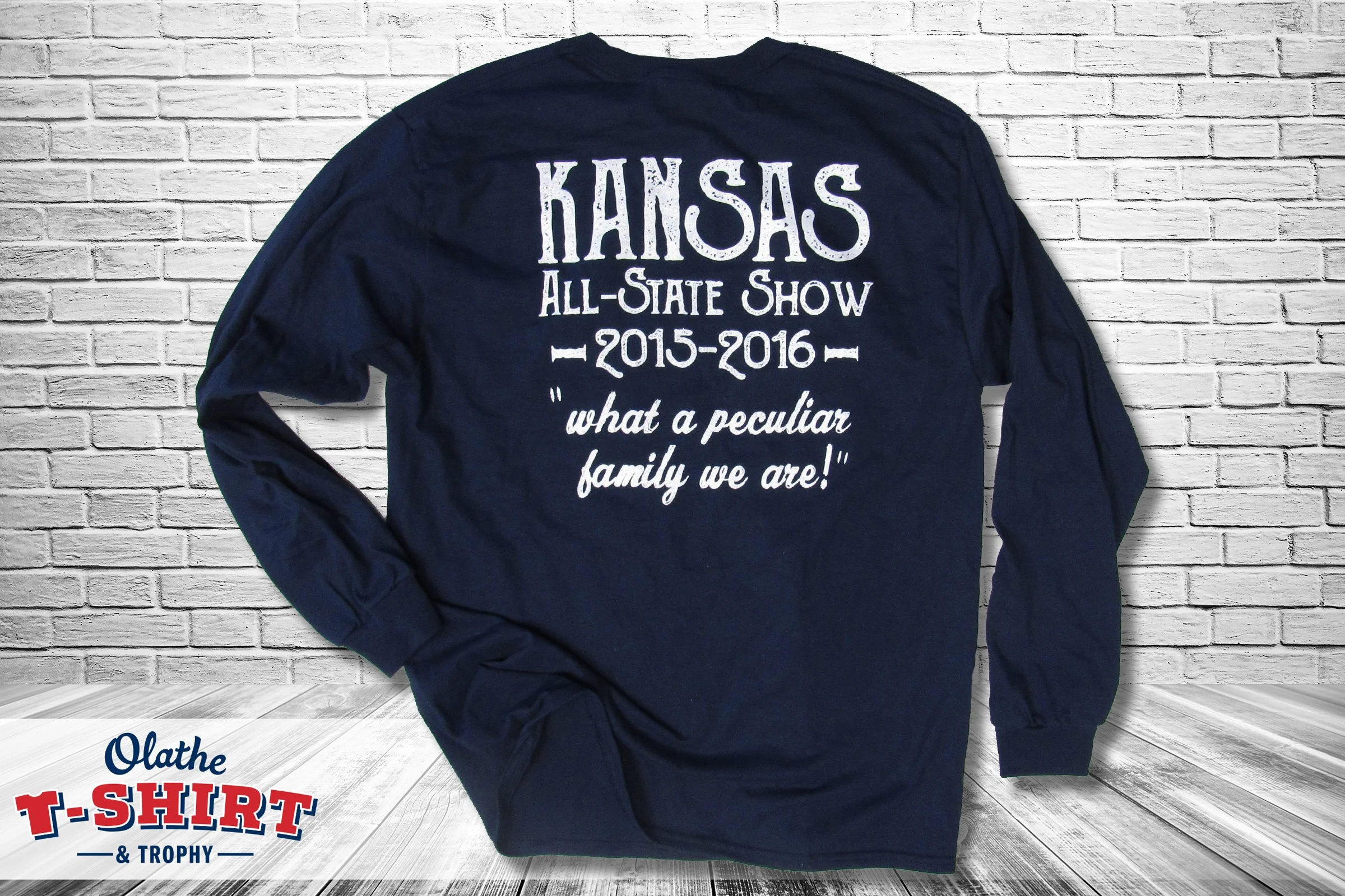 A navy blue long-sleeve shirt hanging against a white brick wall, with white text that reads: "KANSAS ALL-STATE SHOW 2015-2016" and a quote, "what a peculiar family we are!"