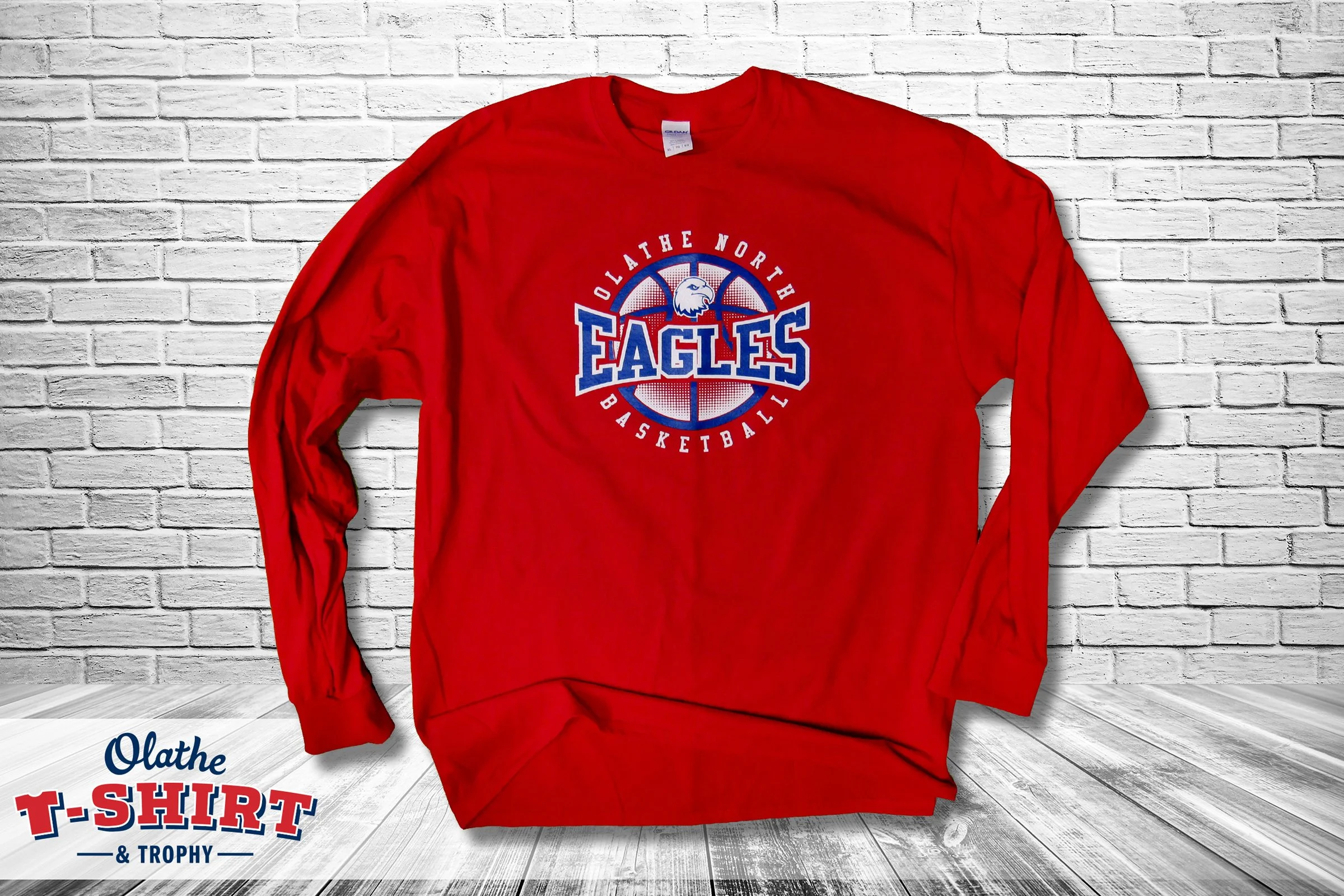 Red long sleeve T-shirt with a basketball logo and the words "Oklahoma North Eagles Basketball" printed on the front, hanging on a white brick wall.